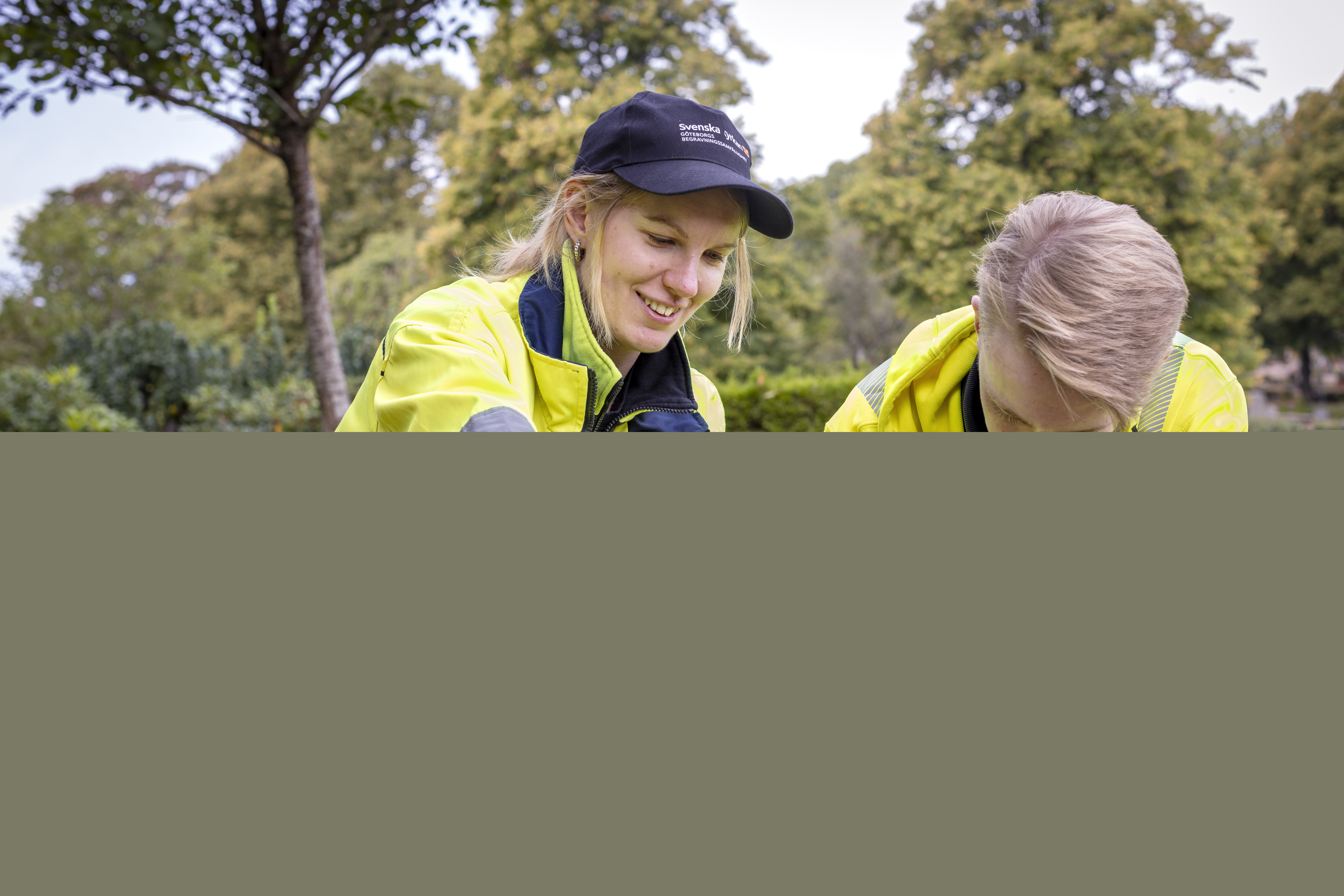 Två kyrkogårdsarbetare i gula varselkläder fixar med några blommor.