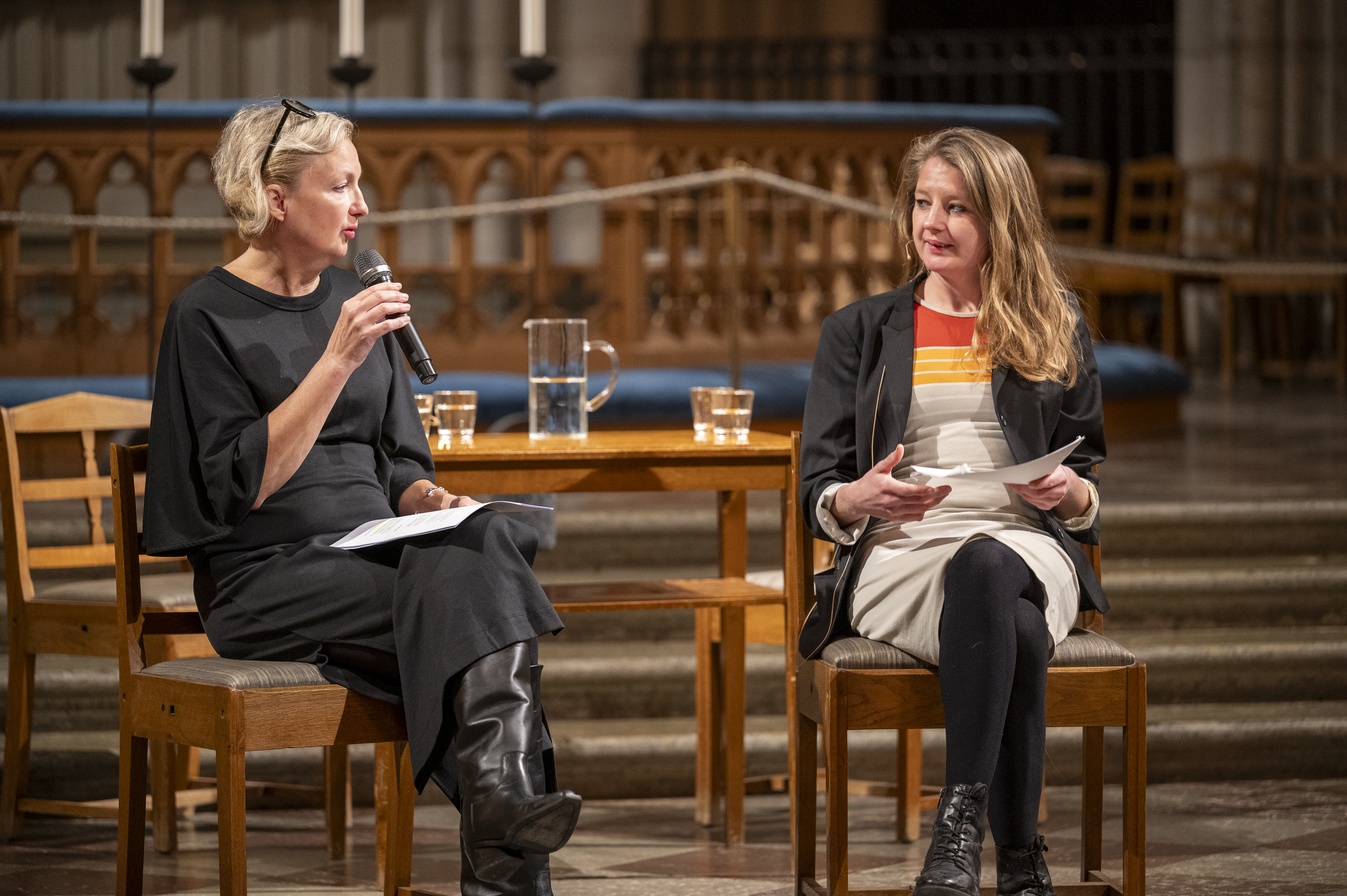 En kvinna med mikrofon i handen sitter vid ett bord i en kyrka och samtalar med en person som sitter bredvid.