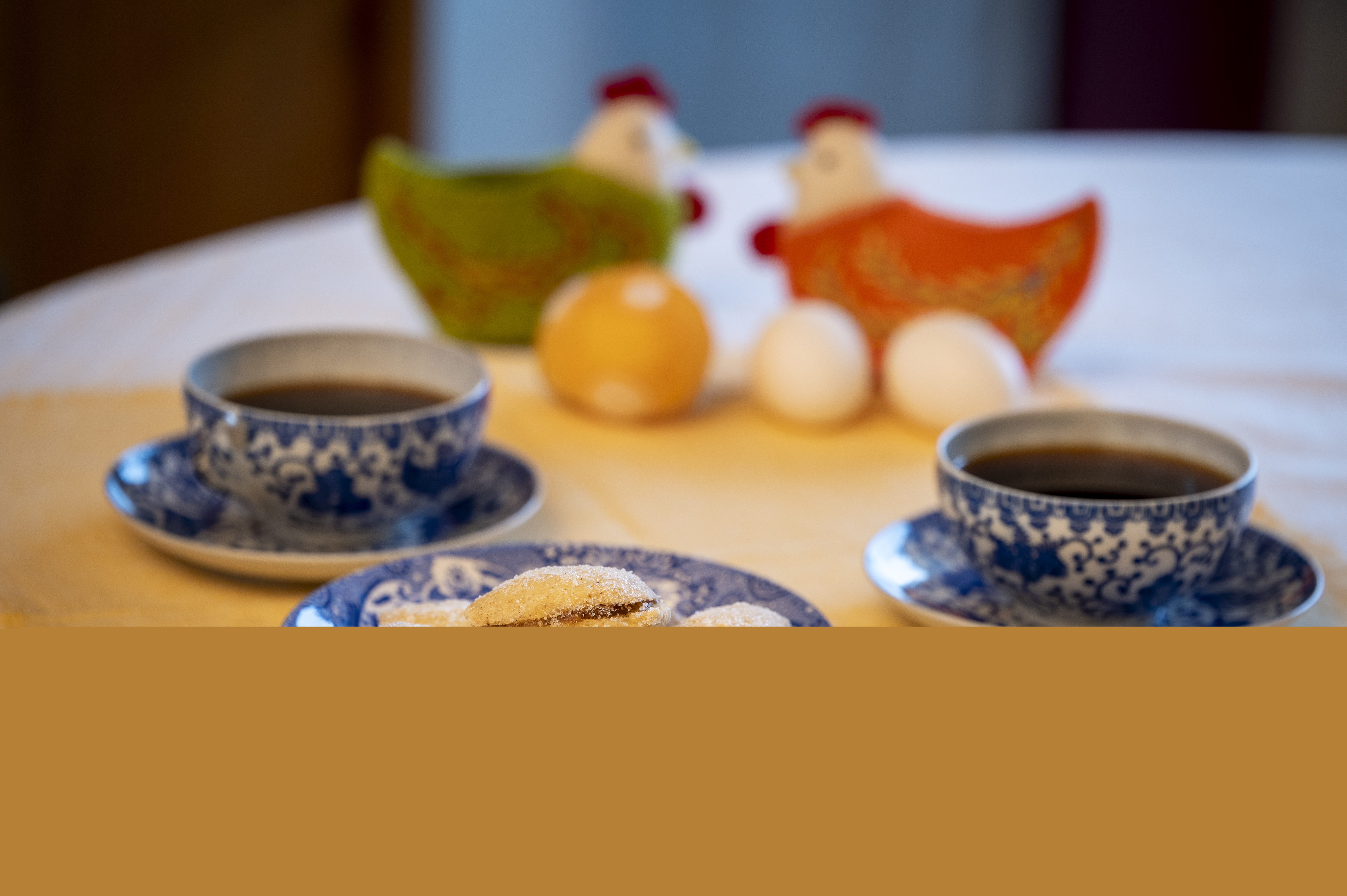 Ett kakfat, två kaffekoppar på fat och två påskhönor med ägg står uppdukade på en gul duk.