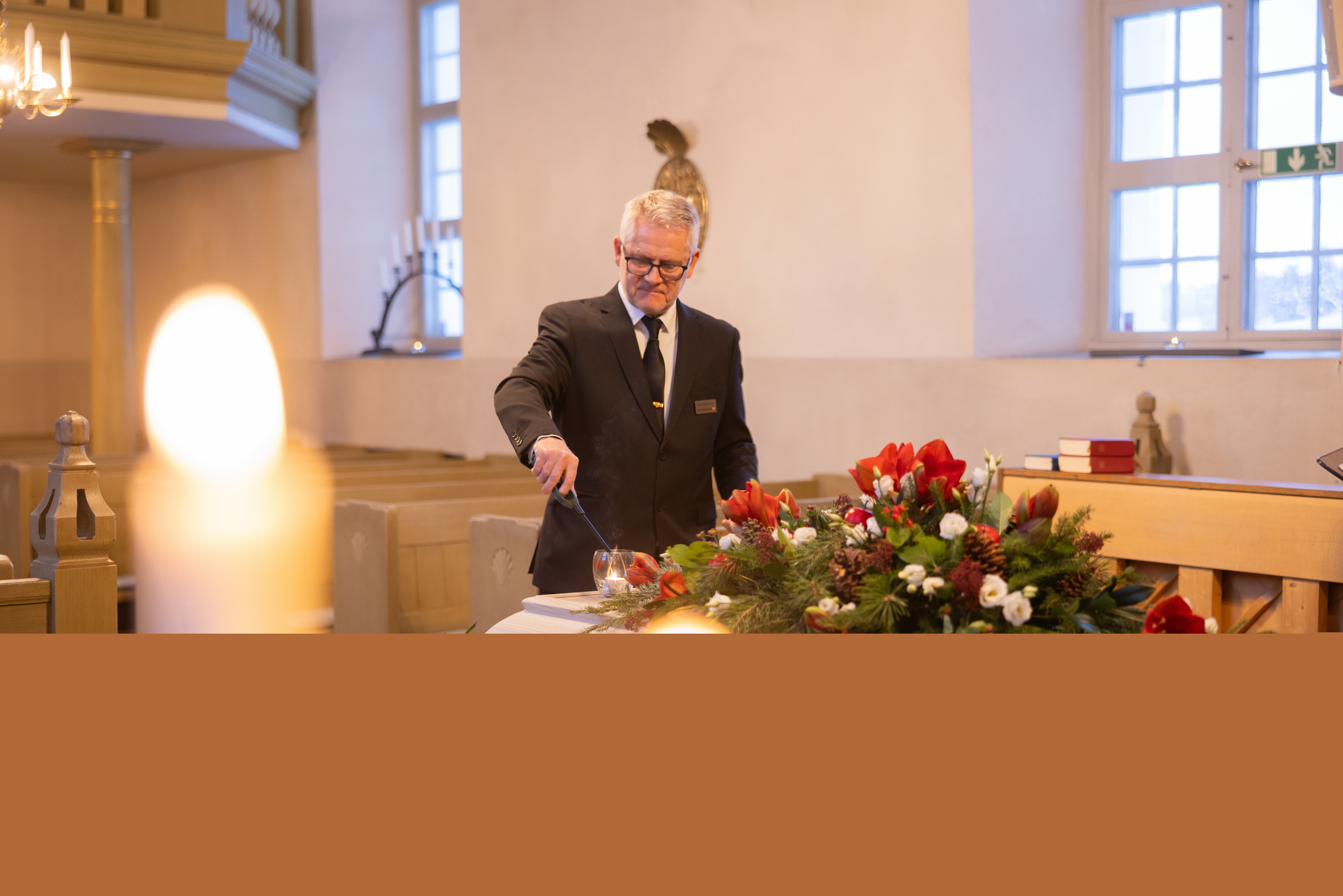 En kyrkvaktmästare tänder ett ljus på en vit kista som har en krans av amaryllis, tallkvistar och vita rosor.