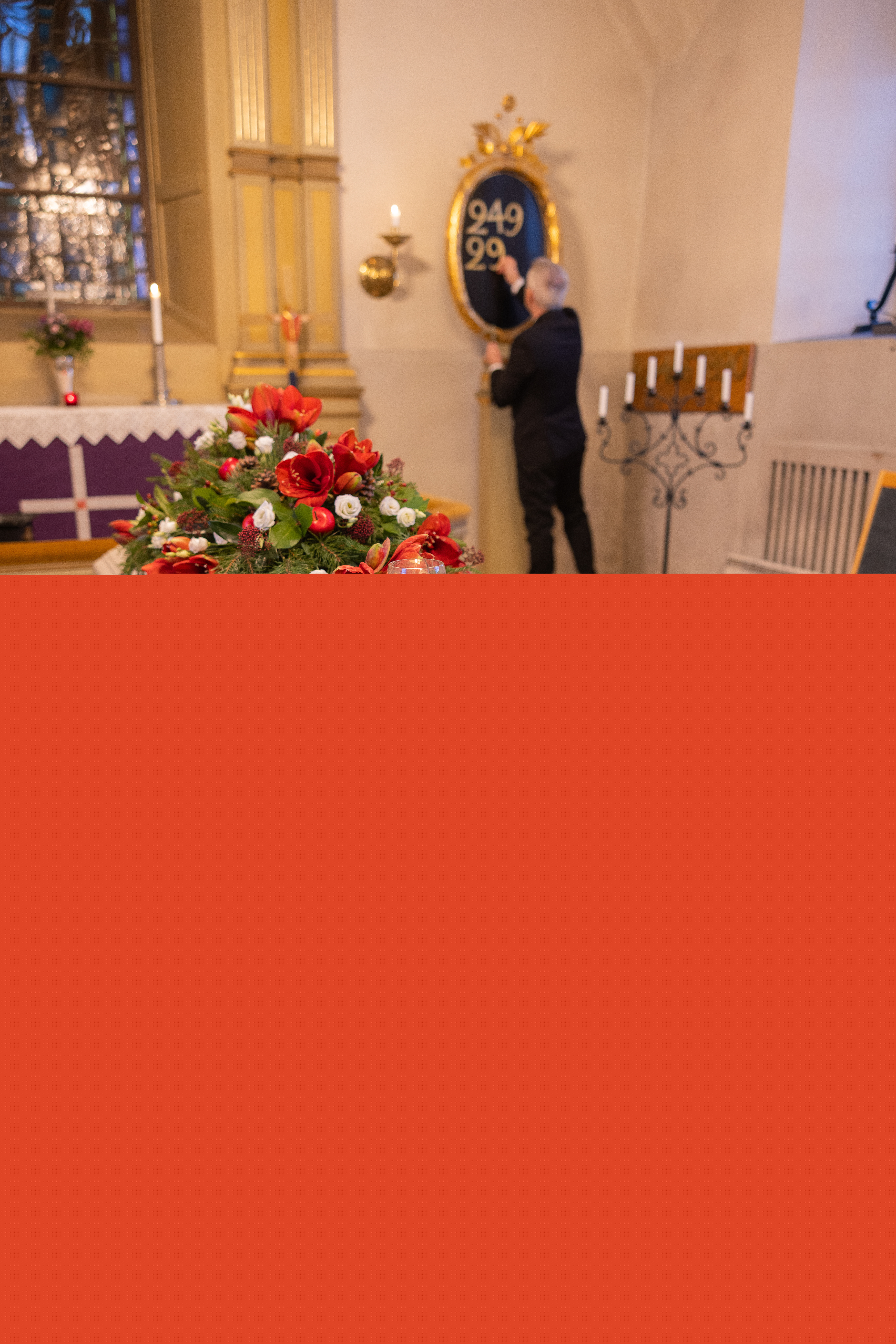 En kyrkvaktmästare hänger upp siffror på psalmtavlan. I koret står en vit kista med röda och vita blommor på.