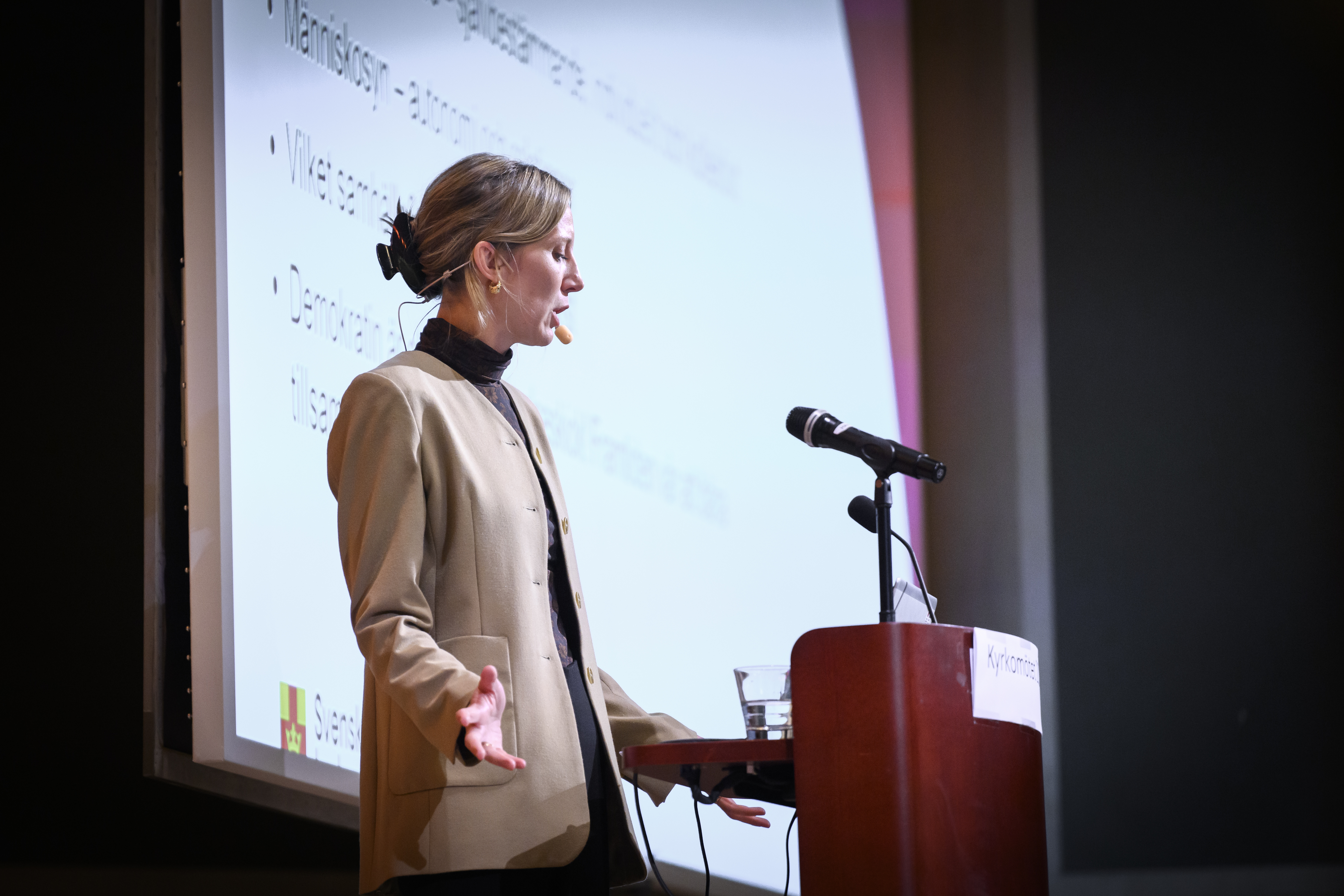 En kvinna med headset står i en talarstol. Hennes presentation visas på storbildsskärm.