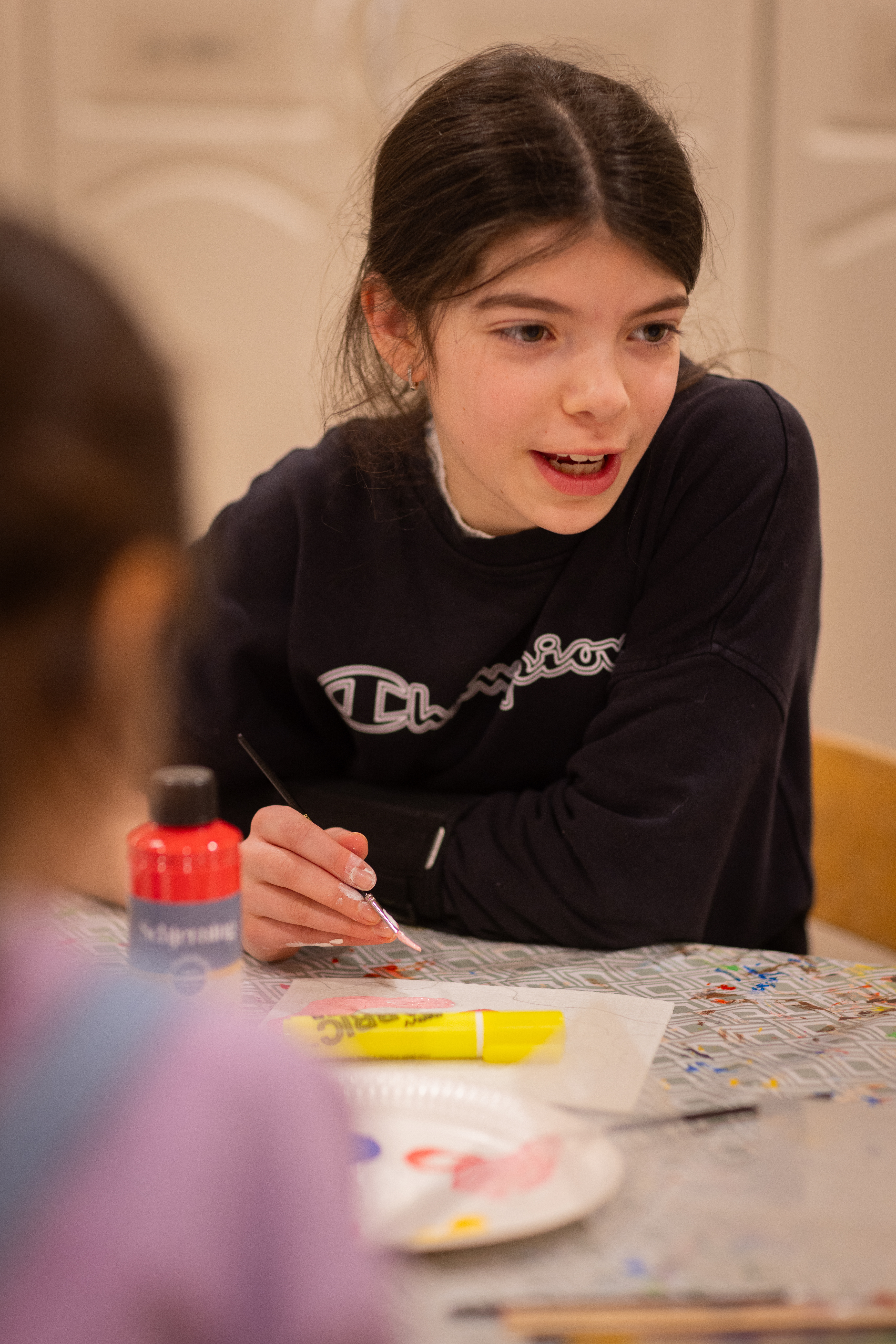En flicka sitter vid ett bord och målar med pensel. Ett annat barn sitter i förgrunden.