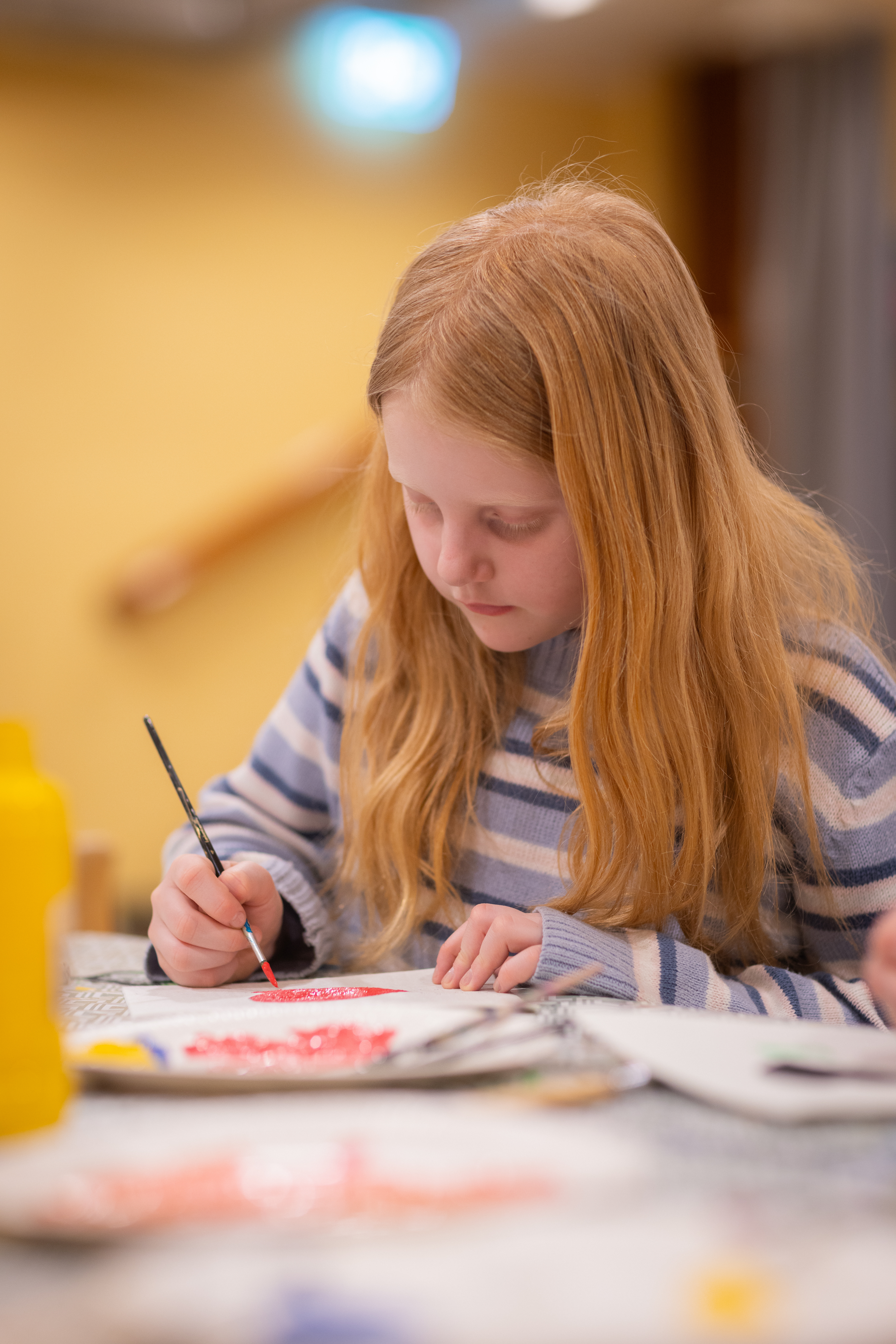 En rödhårig flicka sitter vid ett bord och målar med pensel.