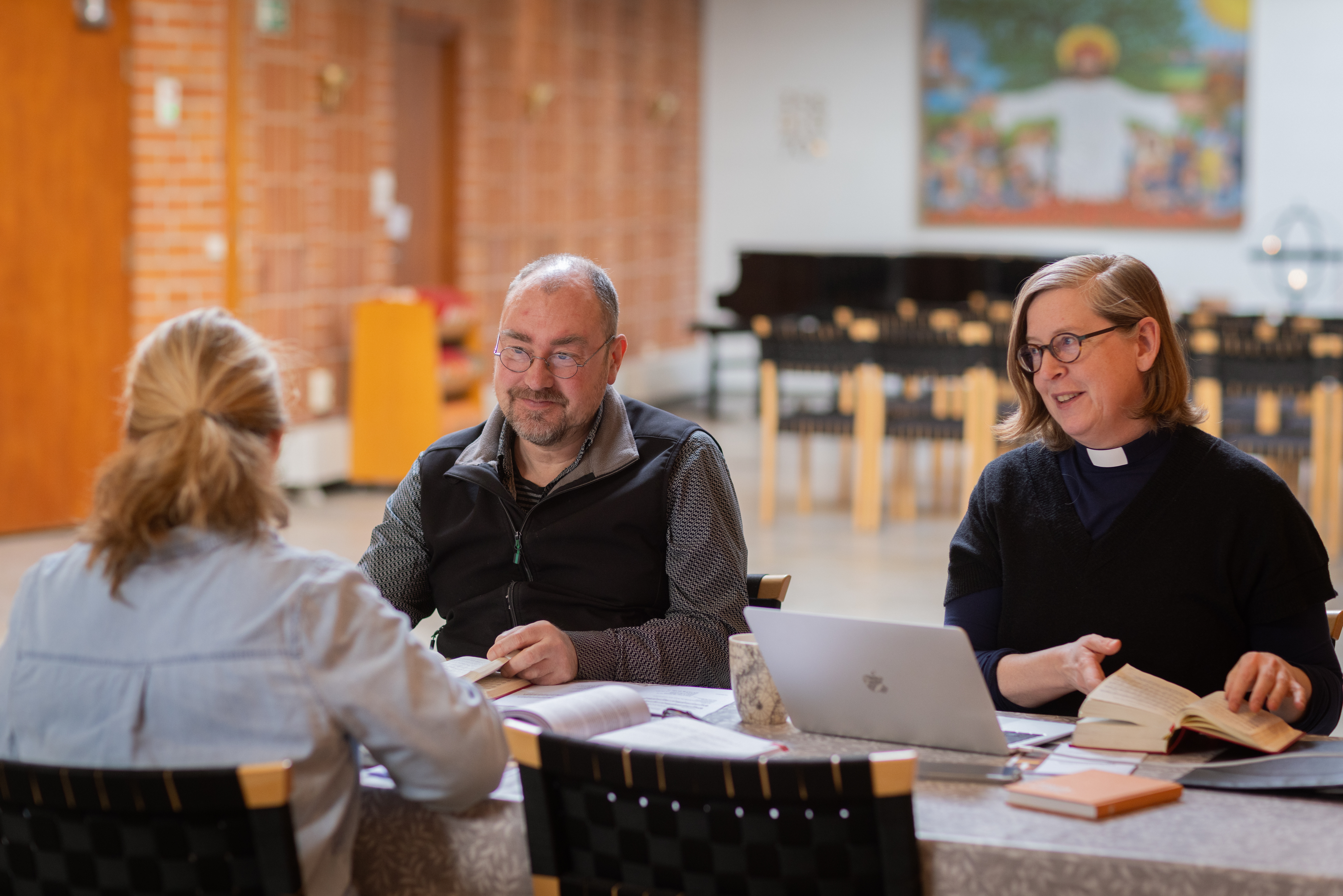 En präst sitter vid ett bord och samtalar med två andra personer.