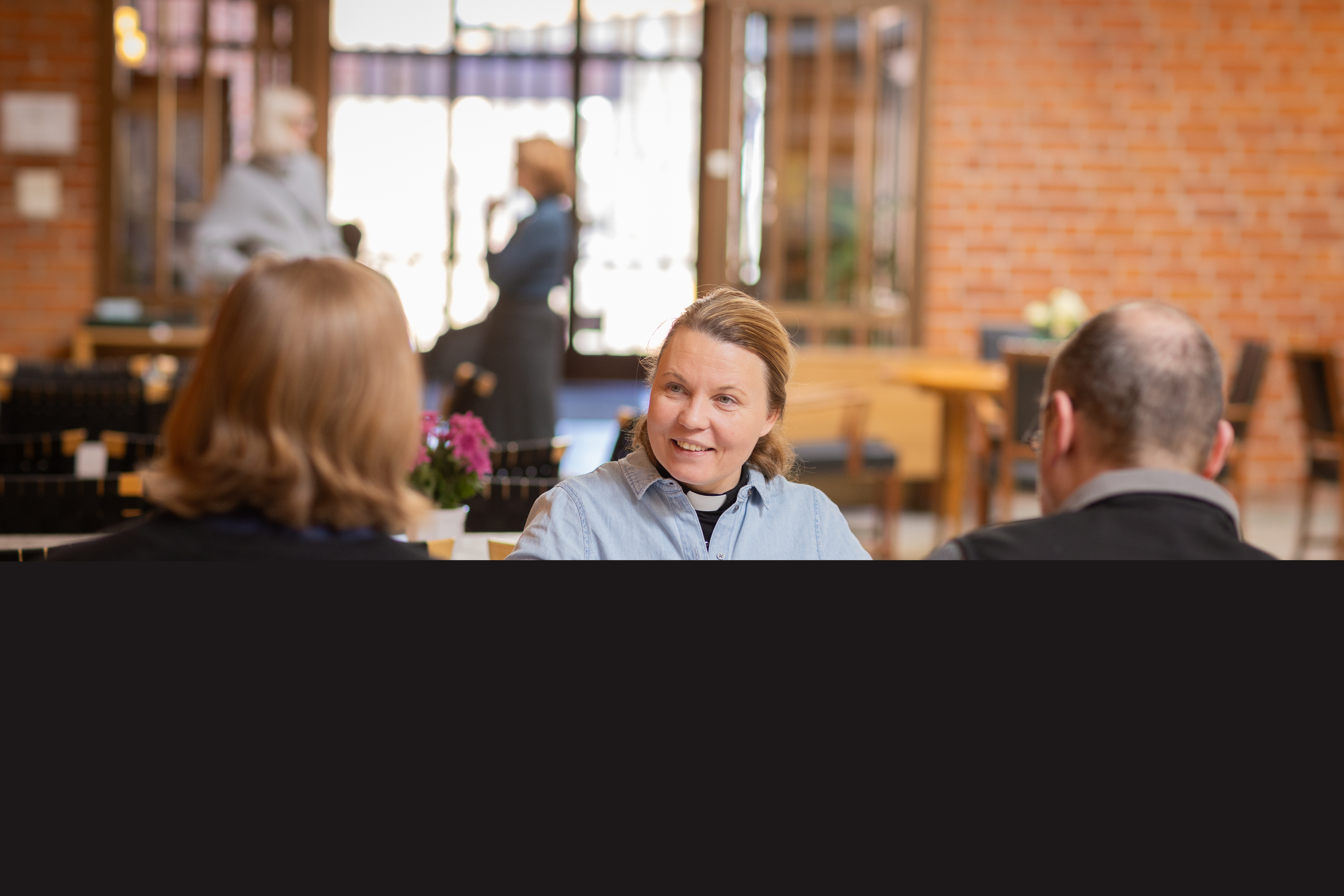 En kvinnlig präst sitter vid ett bord och samtalar med två andra personer.