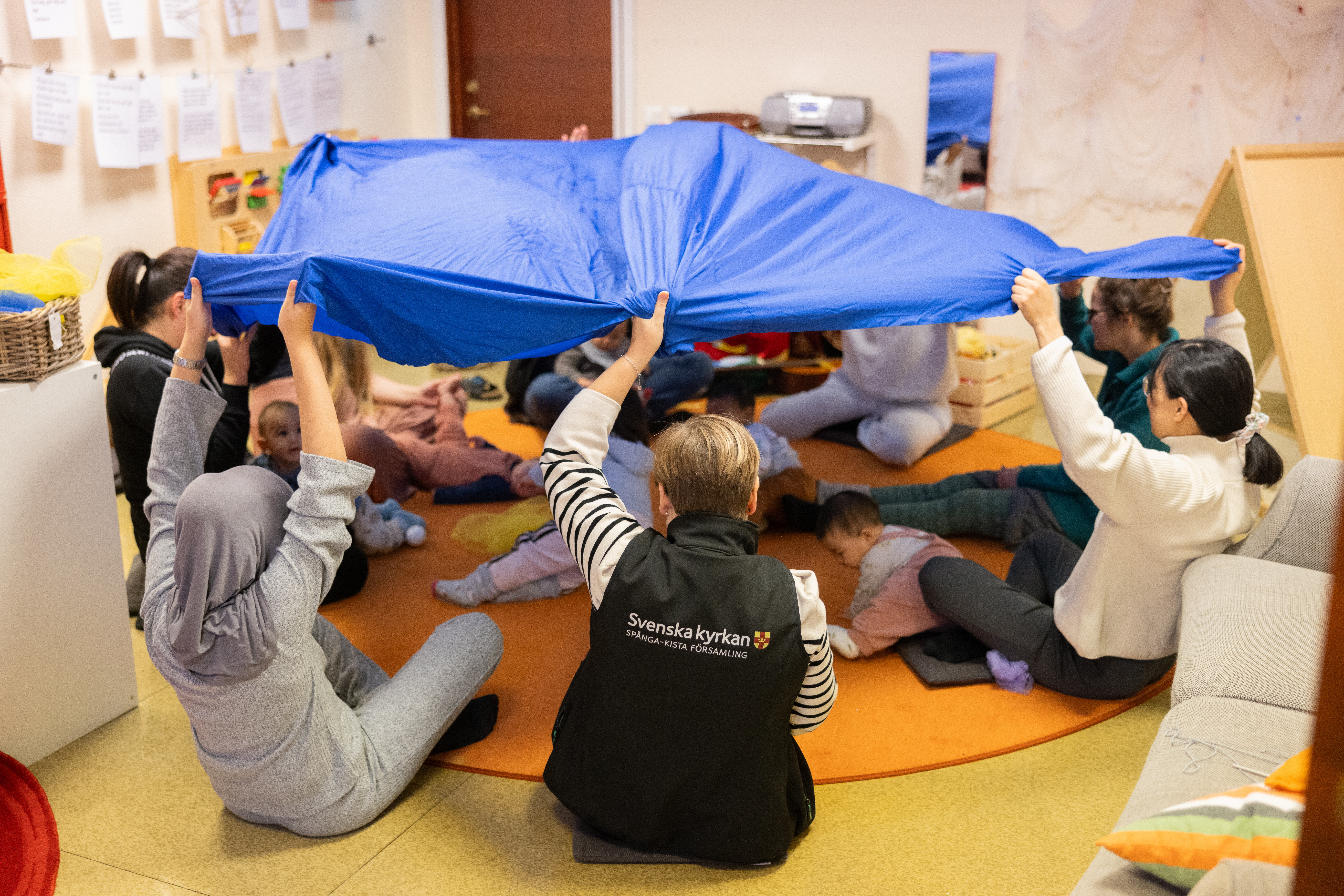 Vuxna personer sitter på golvet i en ring och håller upp ett blått skynke som ett tak över några barn som är därunder.