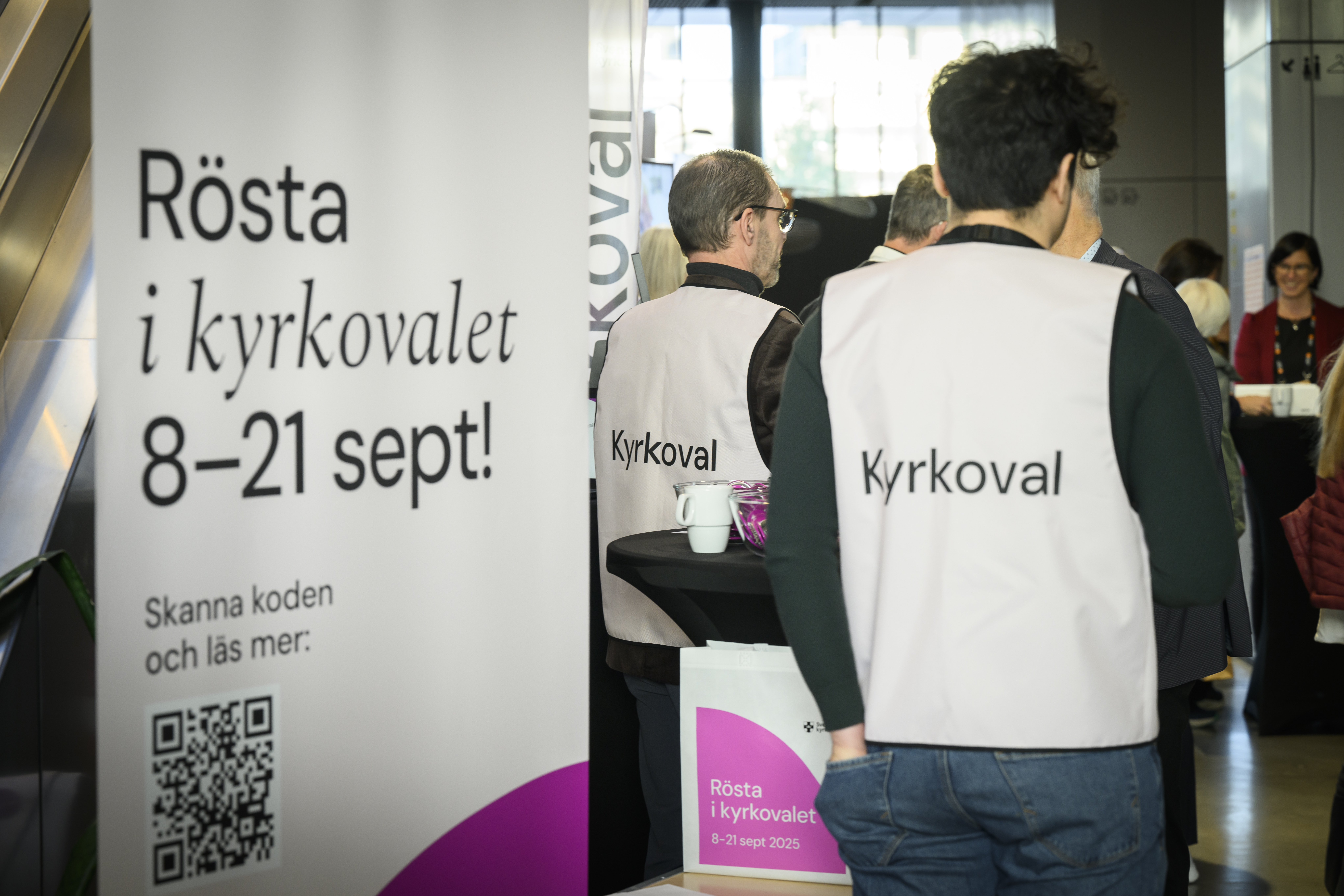 En man i väst med texten Kyrkoval står vid en roll-up med texten Rösta i kyrkovalet 8-21 september.