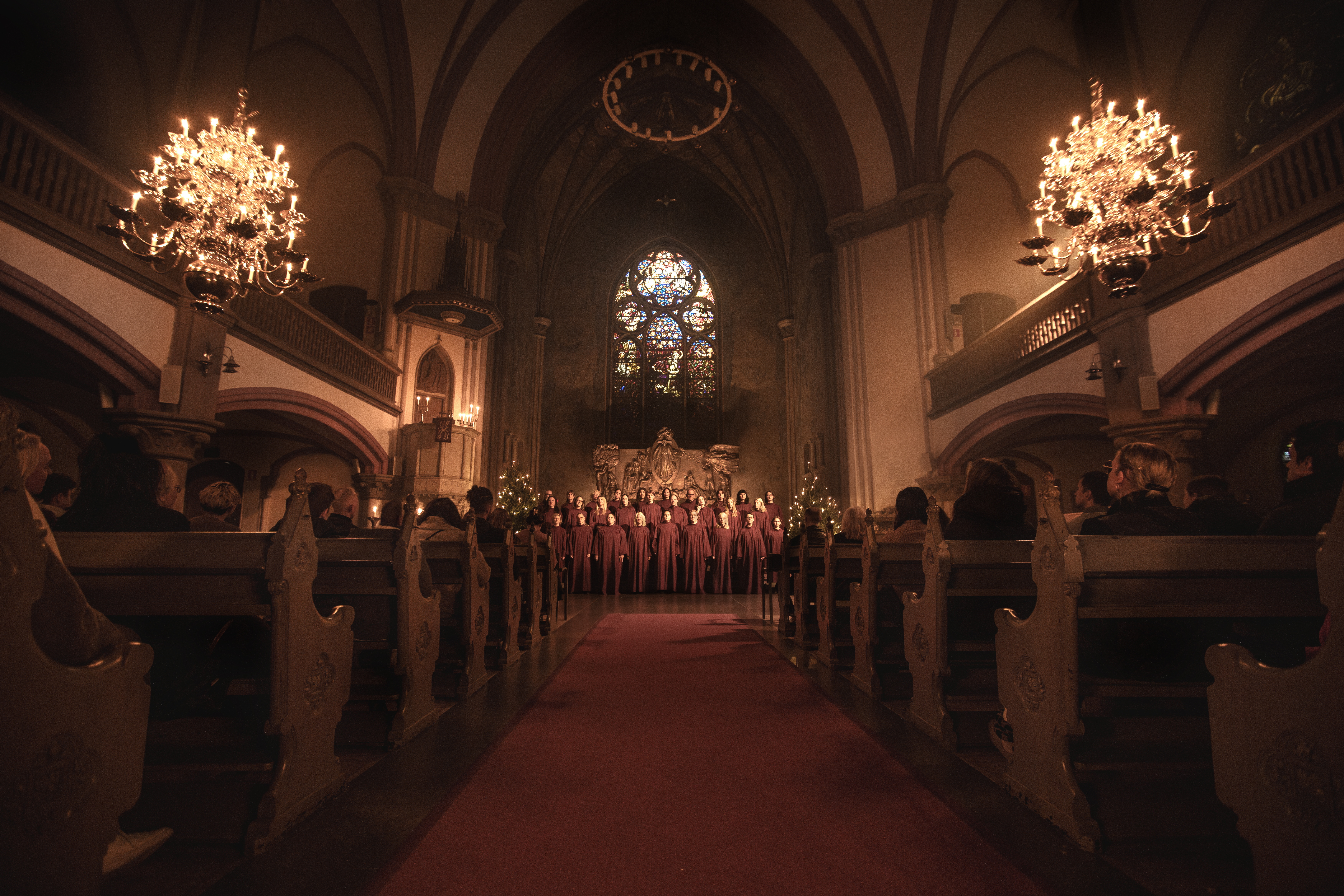 En altargång med kyrkbänkar och två upplysta ljuskronor på varsin sida. En kör står i koret.