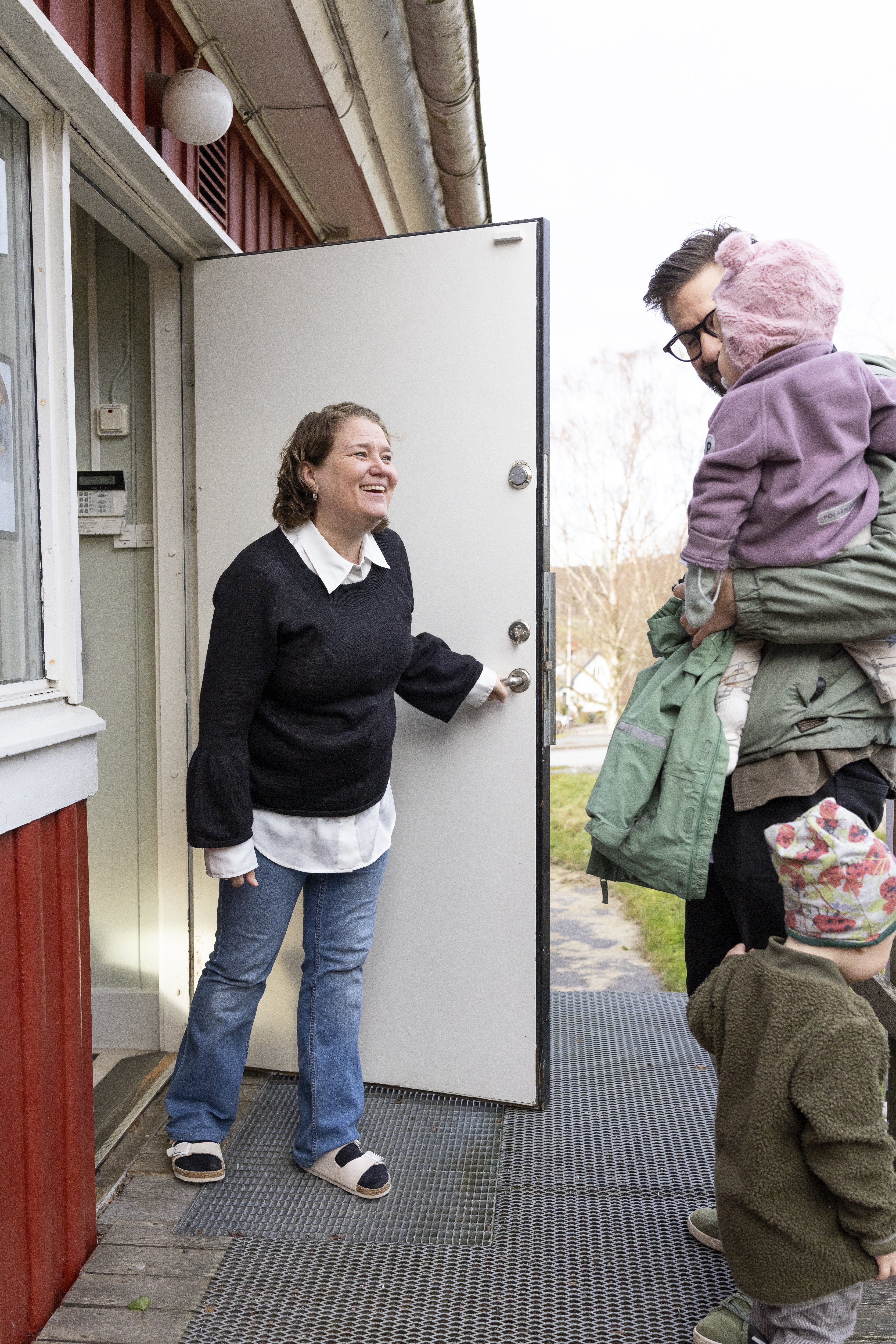 En kvinna öppnar en ytterdörr och hejar på en man och två barn.