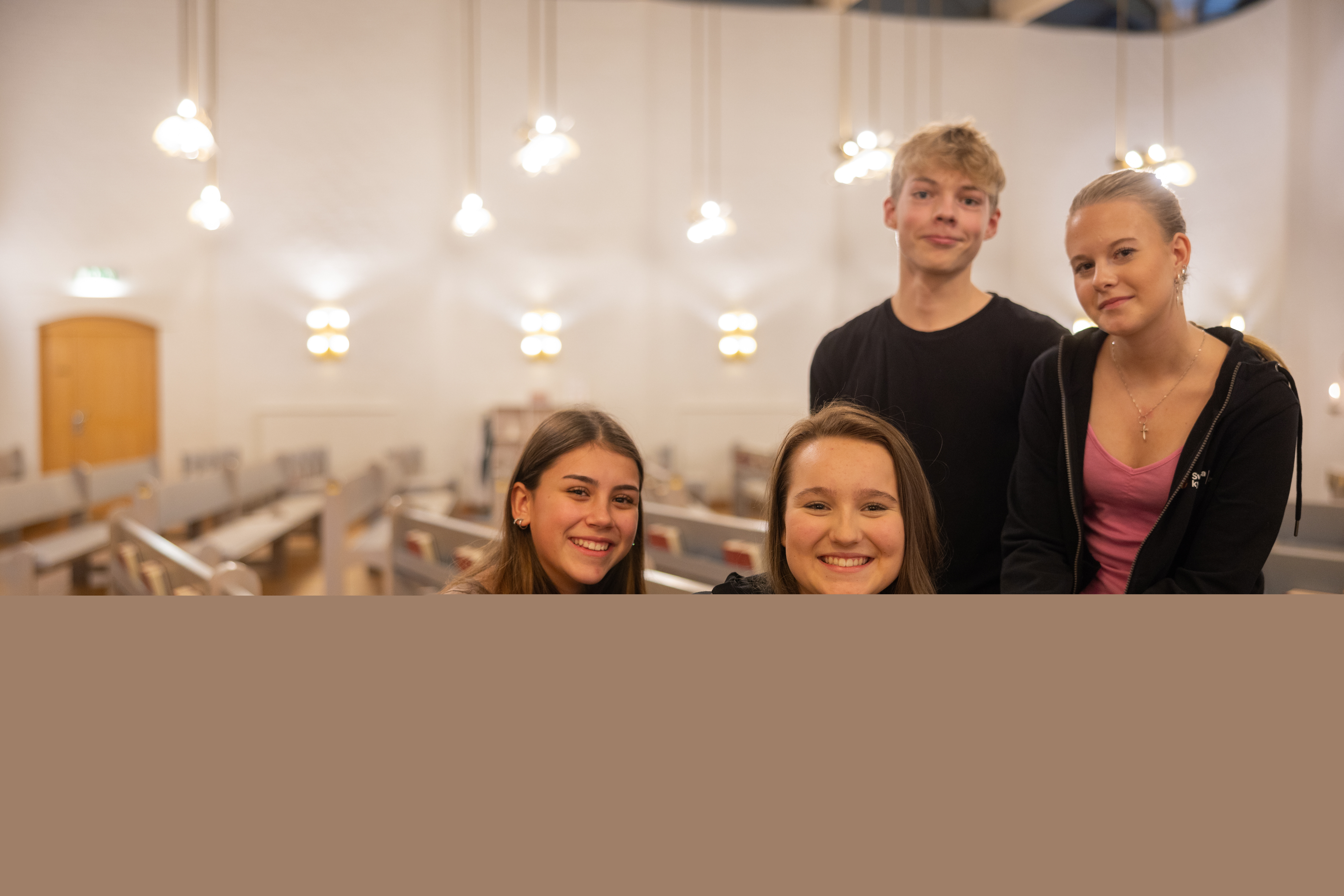 Fyra glada ungdomar sitter i kyrkbänken och tittar in i kameran.