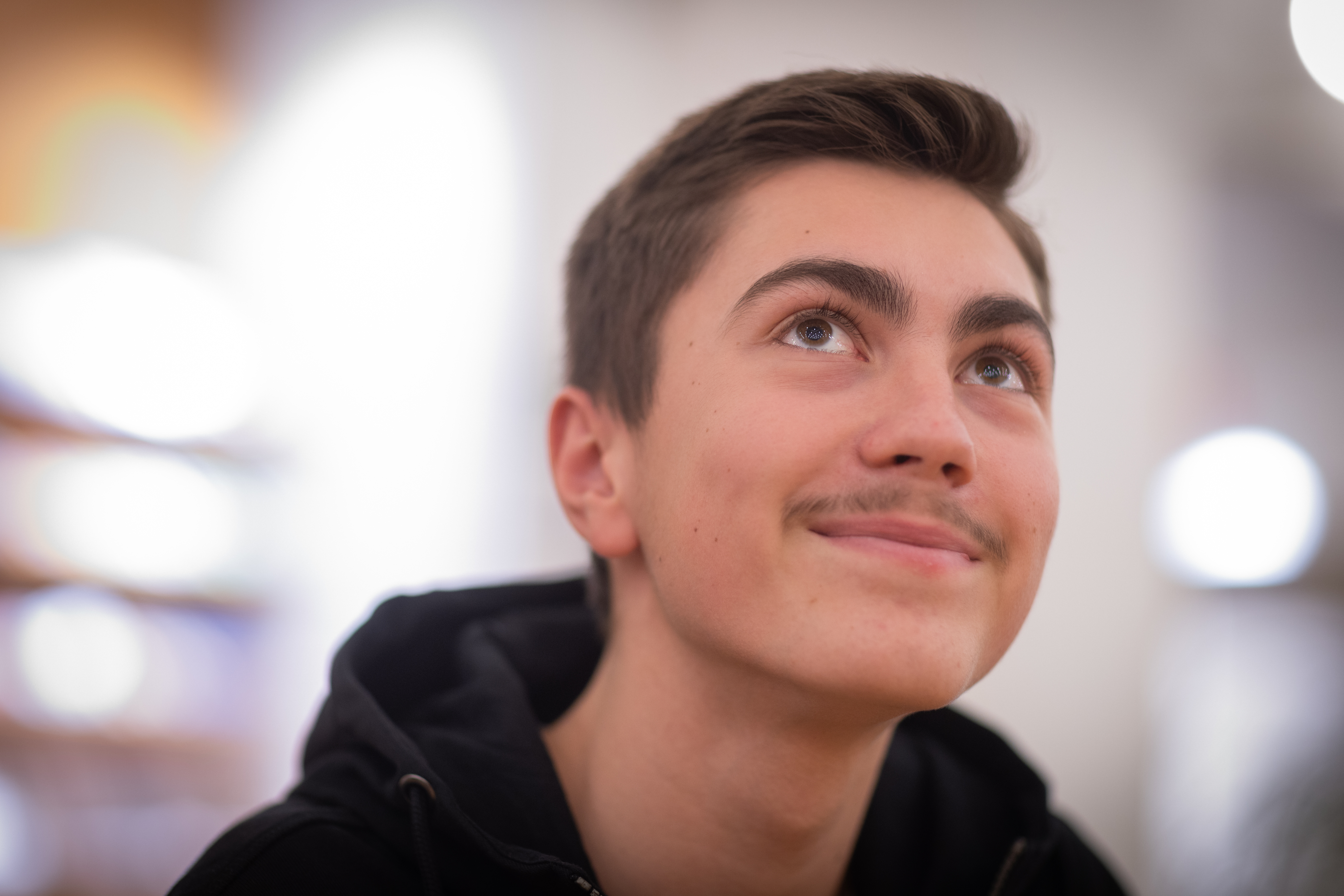 Närbild på en ung leende kille i svart luvtröja som blickar uppåt. Solljus strimmar in från fönstret bakom honom.