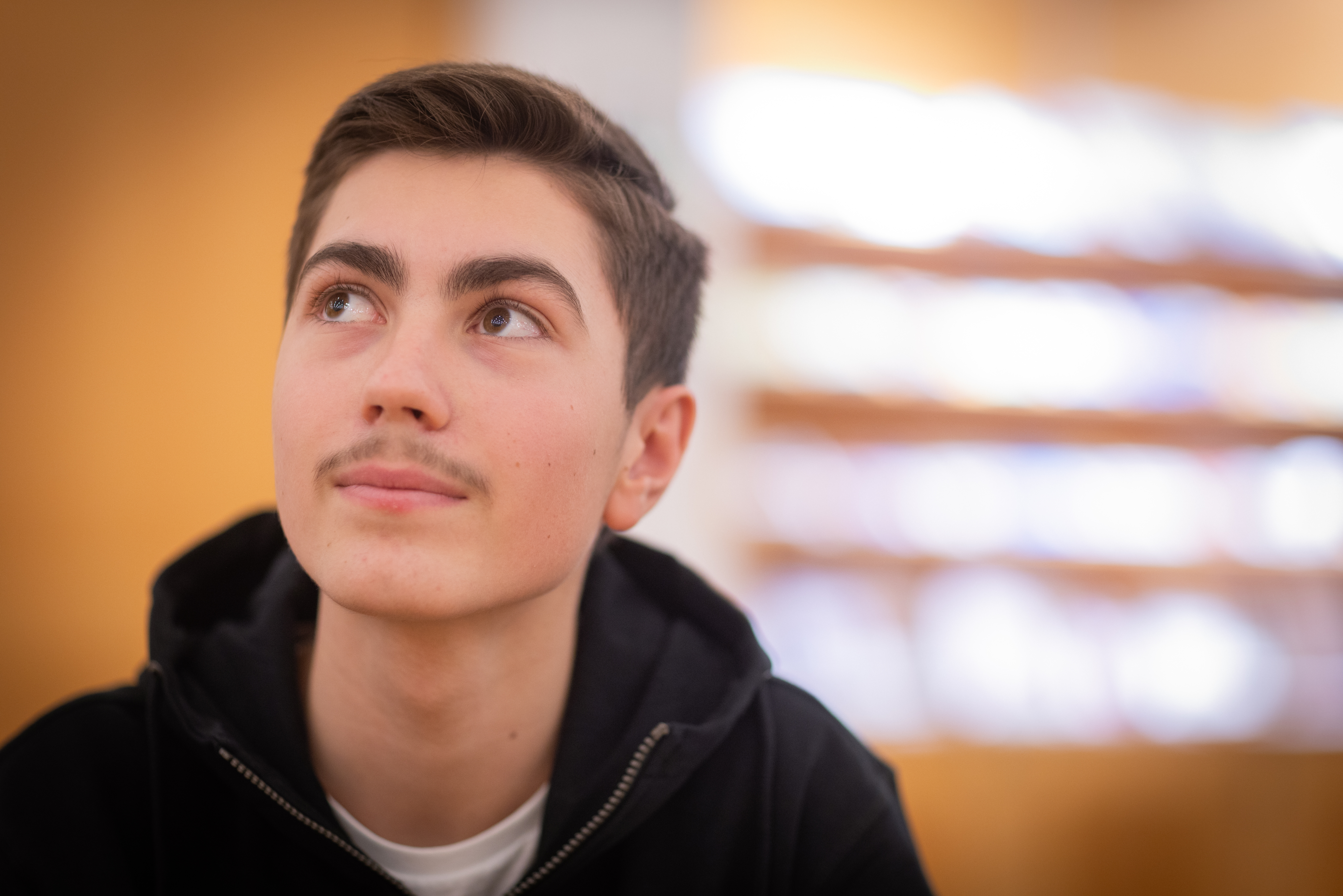 Närbild på en ung kille i svart luvtröja som blickar uppåt. Solljus strimmar in från fönstret bakom honom.
