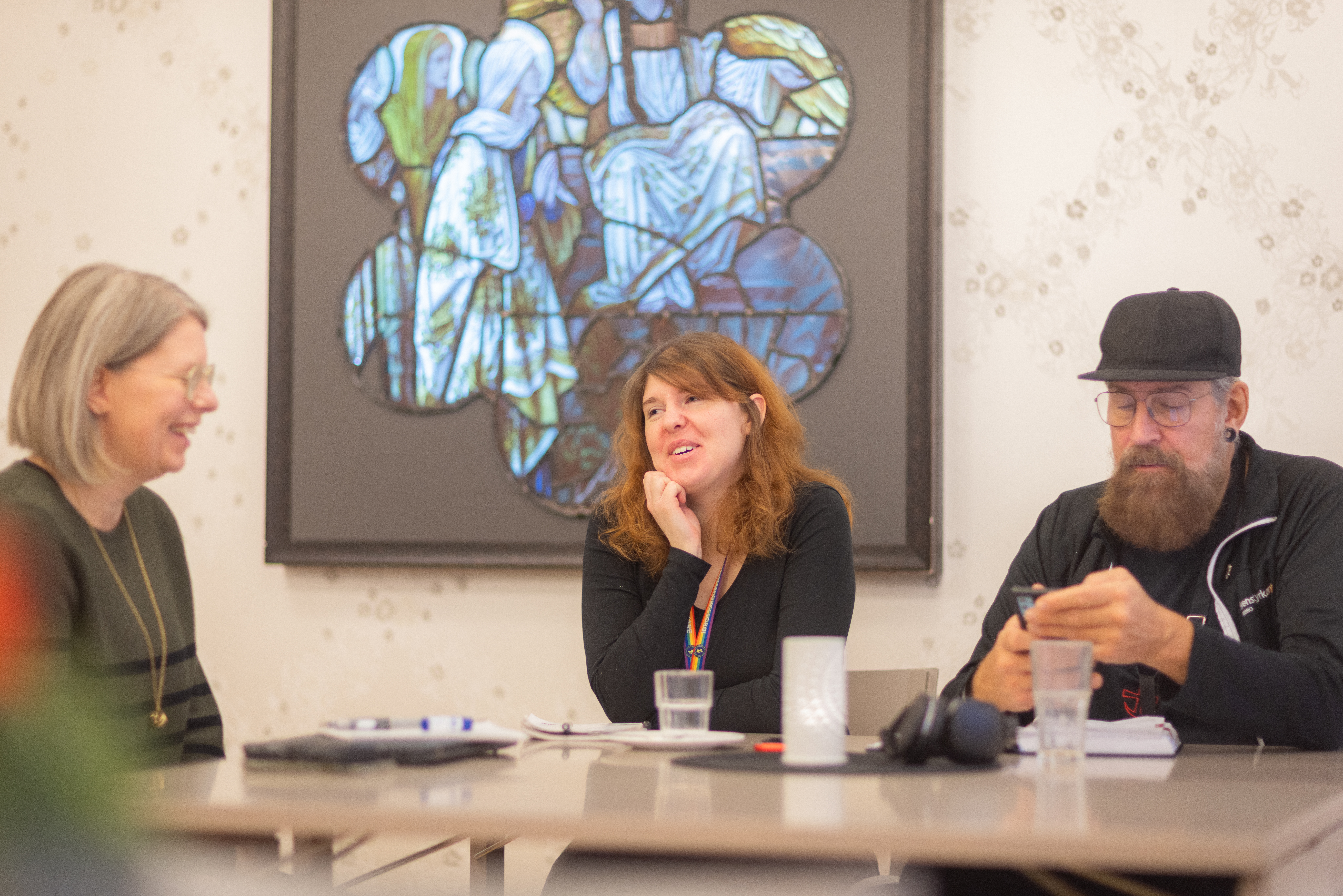 Två kvinnor och en man sitter och samtalar kring ett bord. På väggen bakom hänger en tavla i blå och gröna färger.