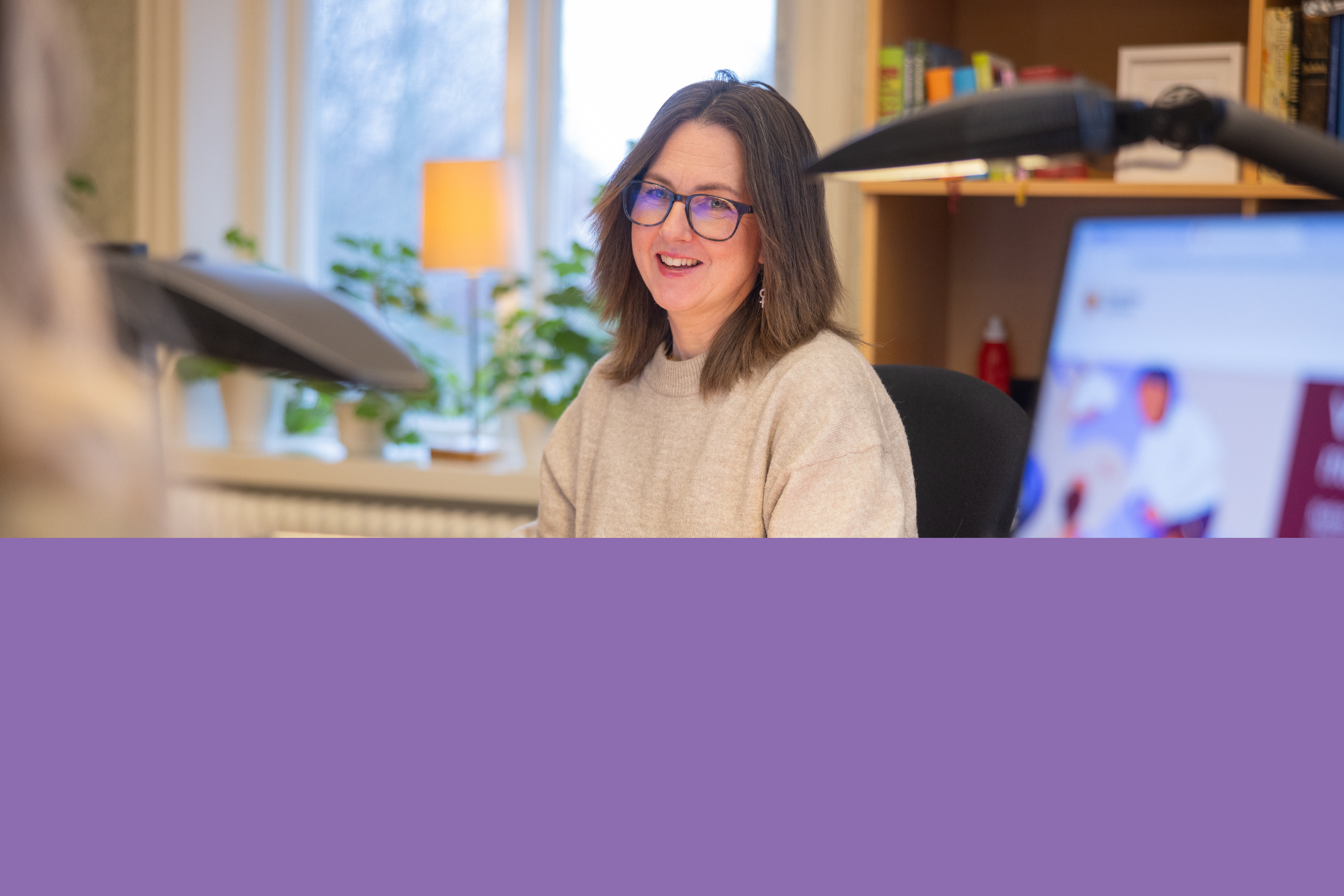 En kvinna i beige tröja sitter och arbetar på ett kontor. Hon pratar med en kollega.