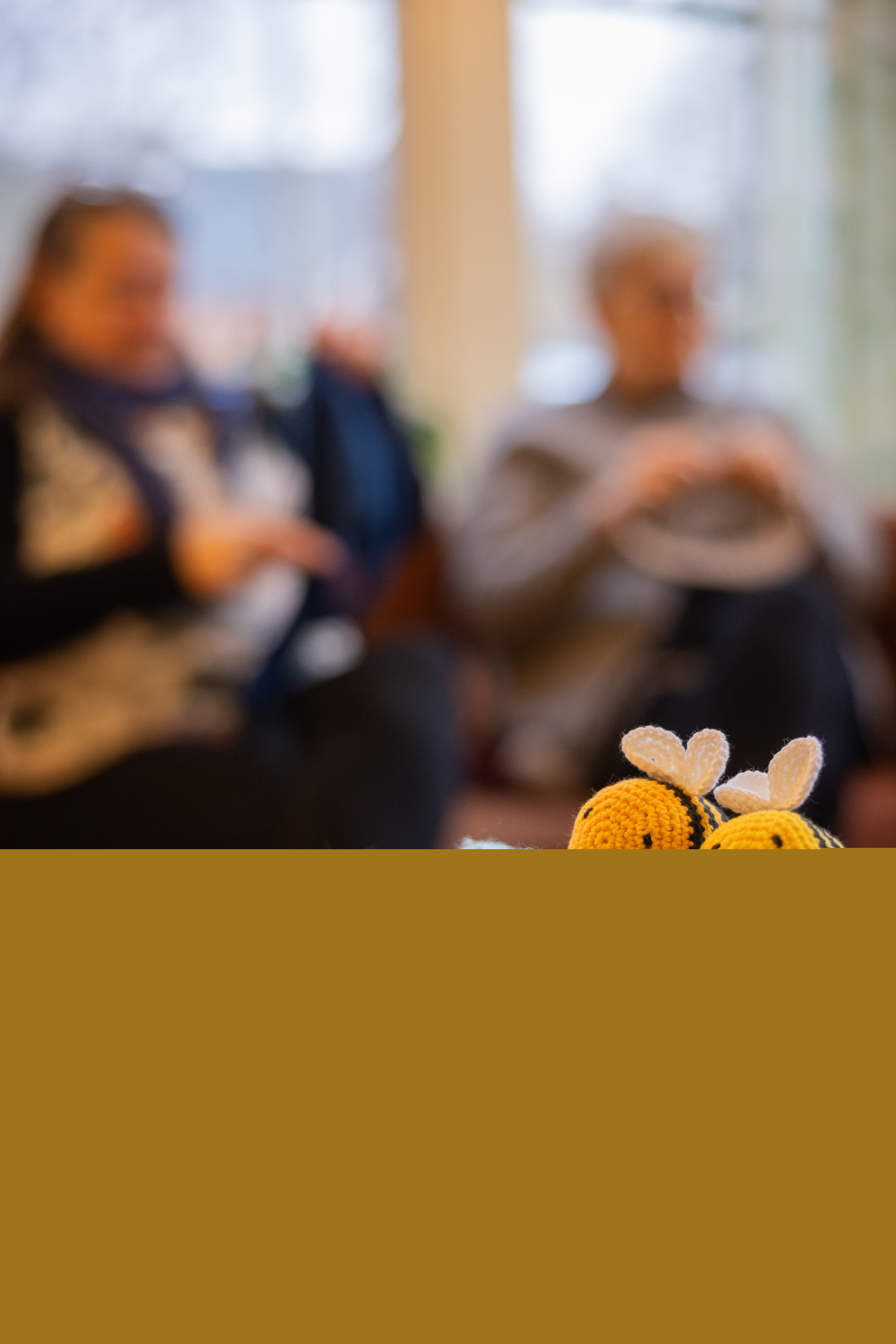 Närbild på garnnystan och två virkade små bin i en korg. Två personer stickar i bakgrunden.