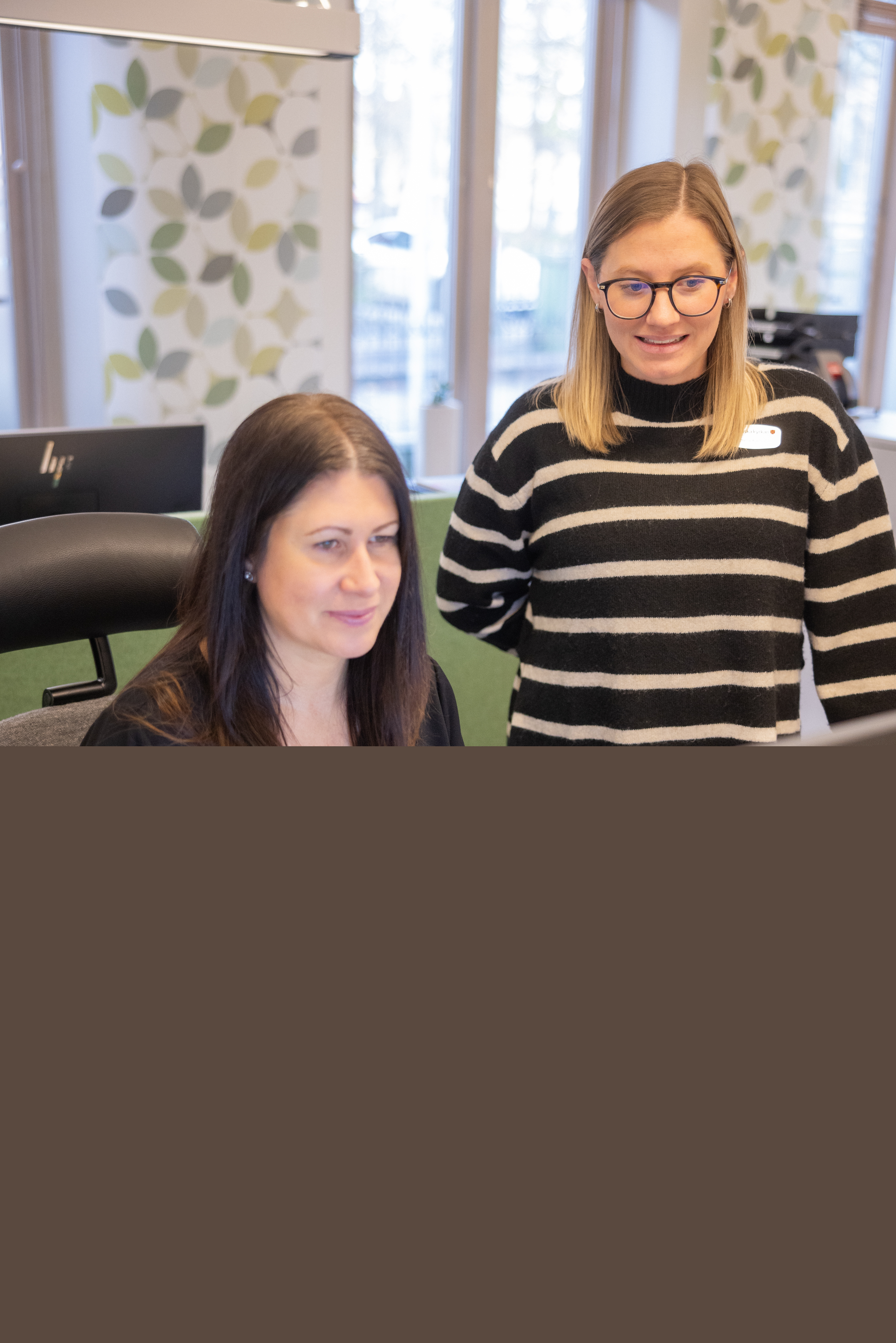 Två kvinnor framför en dator i en reception. Vita gardiner med gröna löv i bakgrunden.