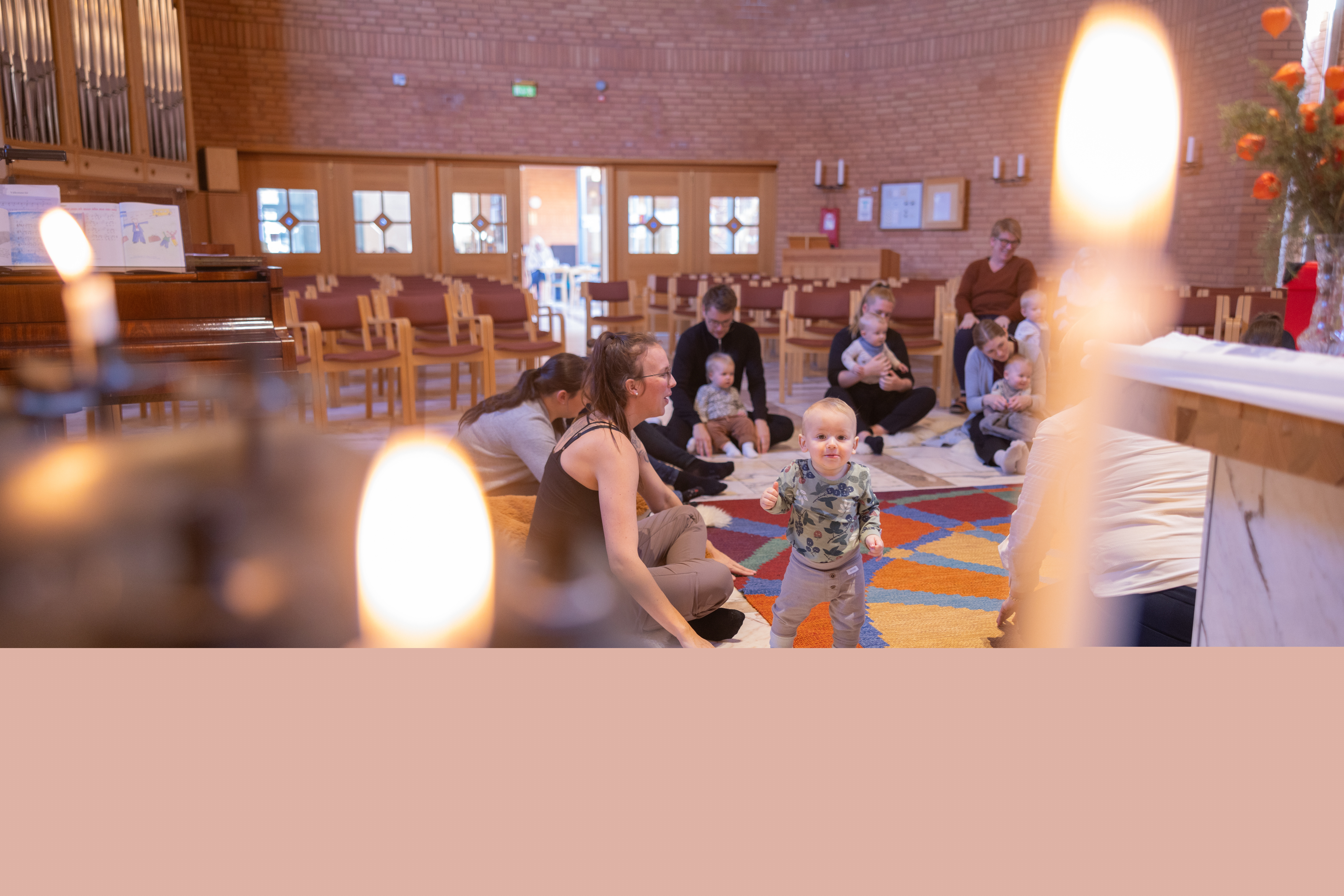 Vuxna och barn sitter på golvet i en kyrka och har sångstund på en öppen förskola. Två tända ljus i förgrunden.