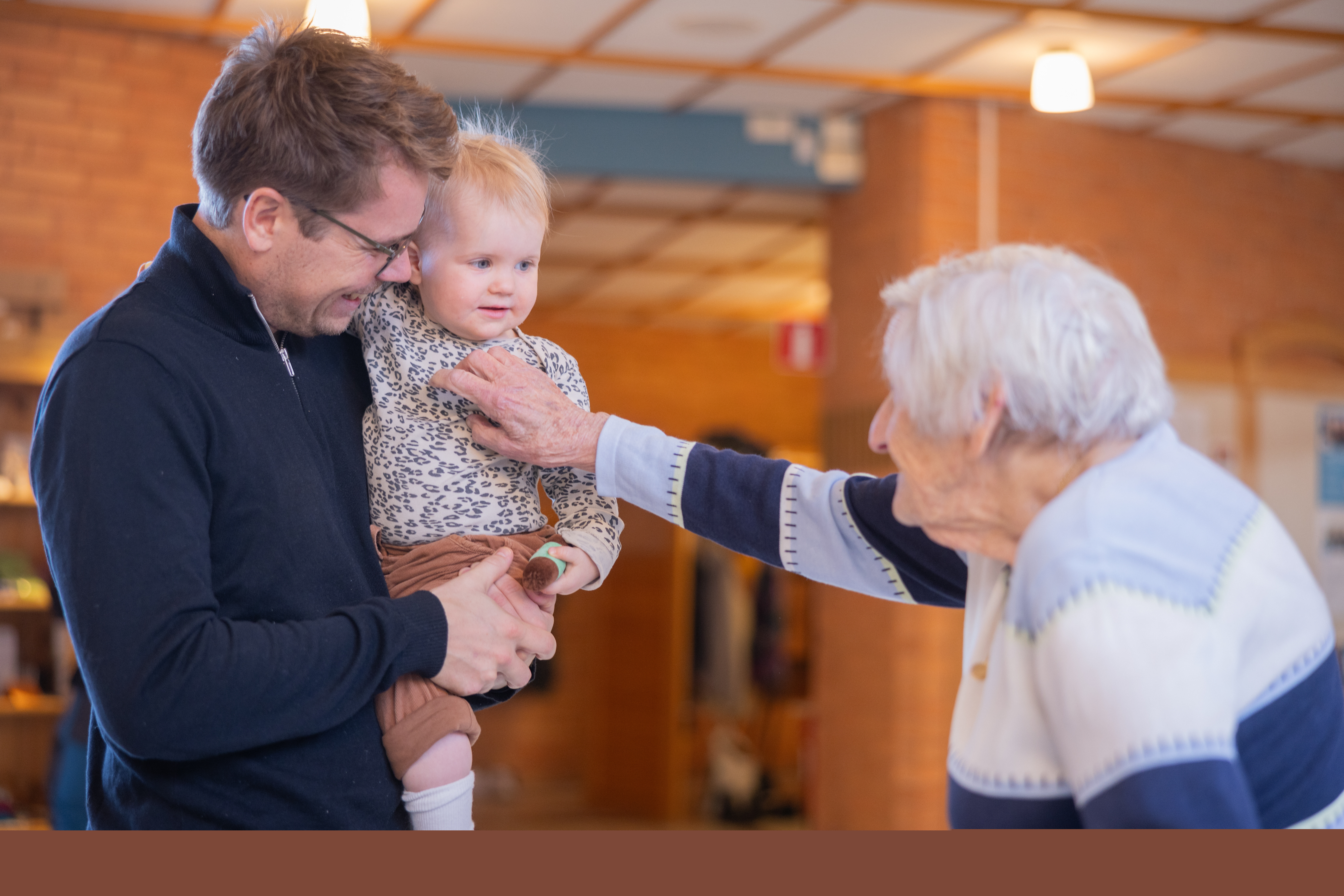 En äldre kvinna busar lekfullt med ett litet barn som sitter i famnen på en vuxen person.