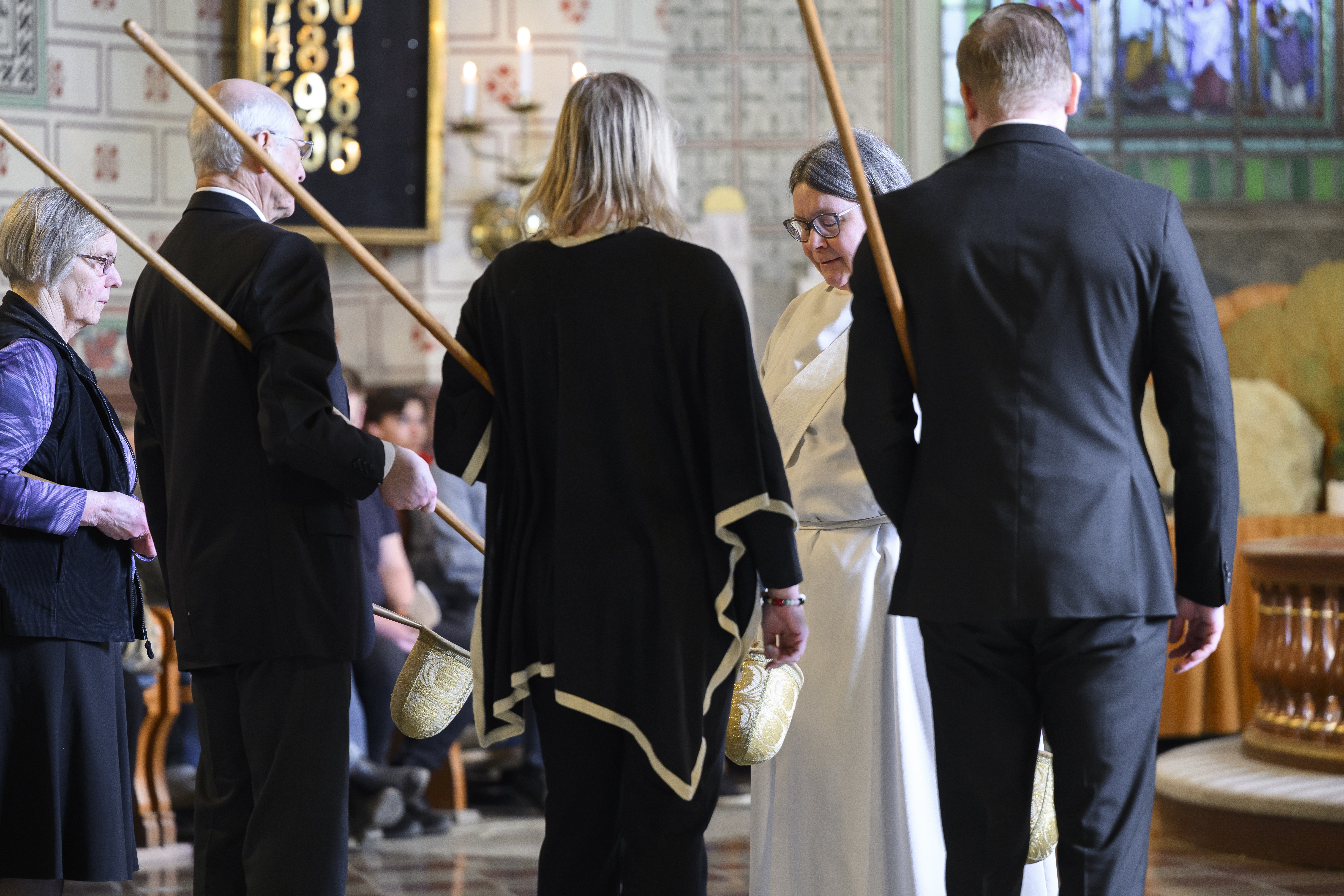 Fyra svartklädda kyrkvärdar står framför en präst med varsin kollekthåv under armen.