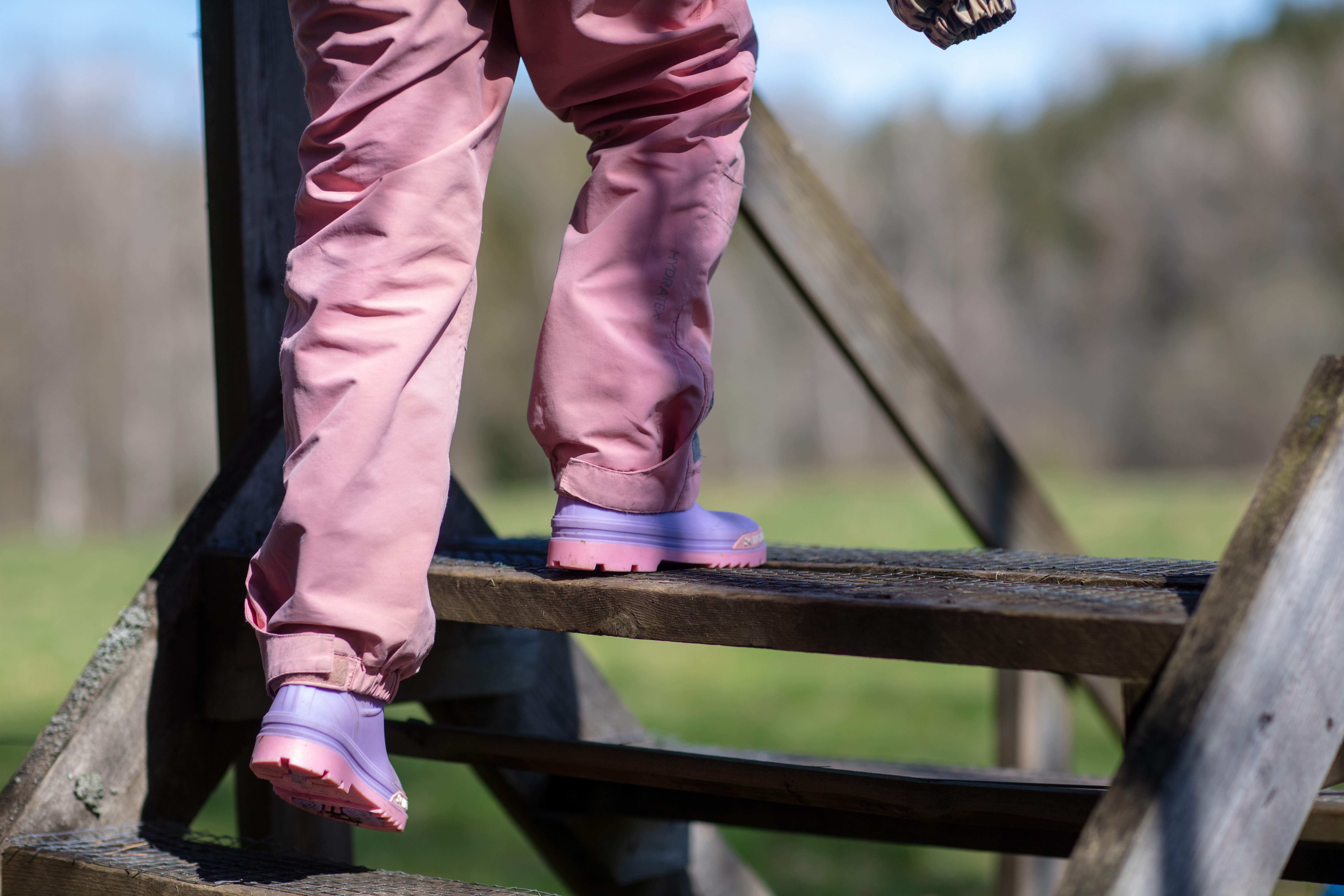 Ett barn i rosa täckbyxor och lila stövlar går över en trappassage vid ett naturreservat.