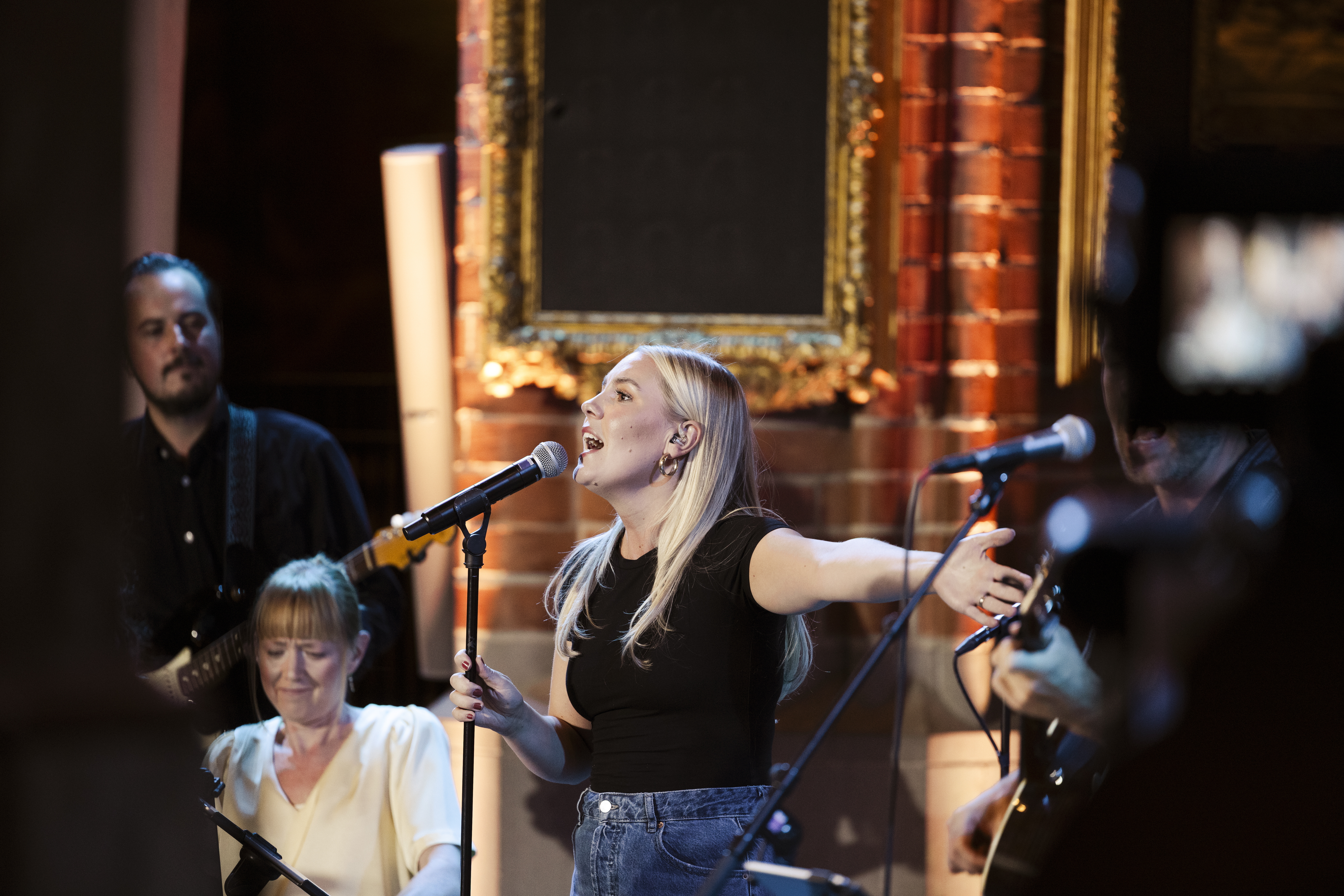 En kvinna står i en kyrka och sjunger i en mikrofon. Runt om henne är det musiker.