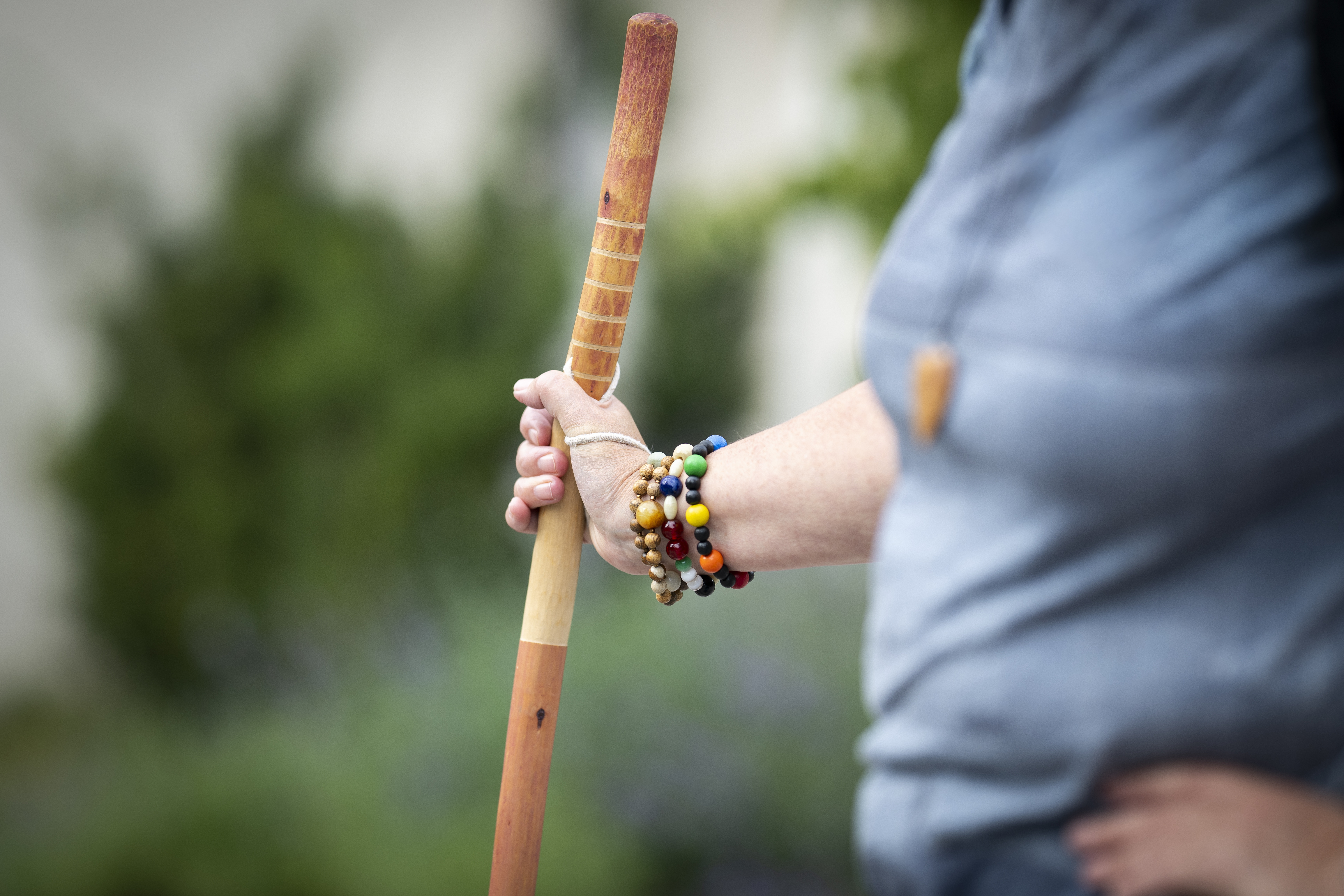 Närbild på en person som håller i en vandringsstav med höger hand. Personen bär flera färgglada armband med pärlor på handleden och har på sig en ljusblå tröja. Bakgrunden är suddig och visar gröna buskar och växter.