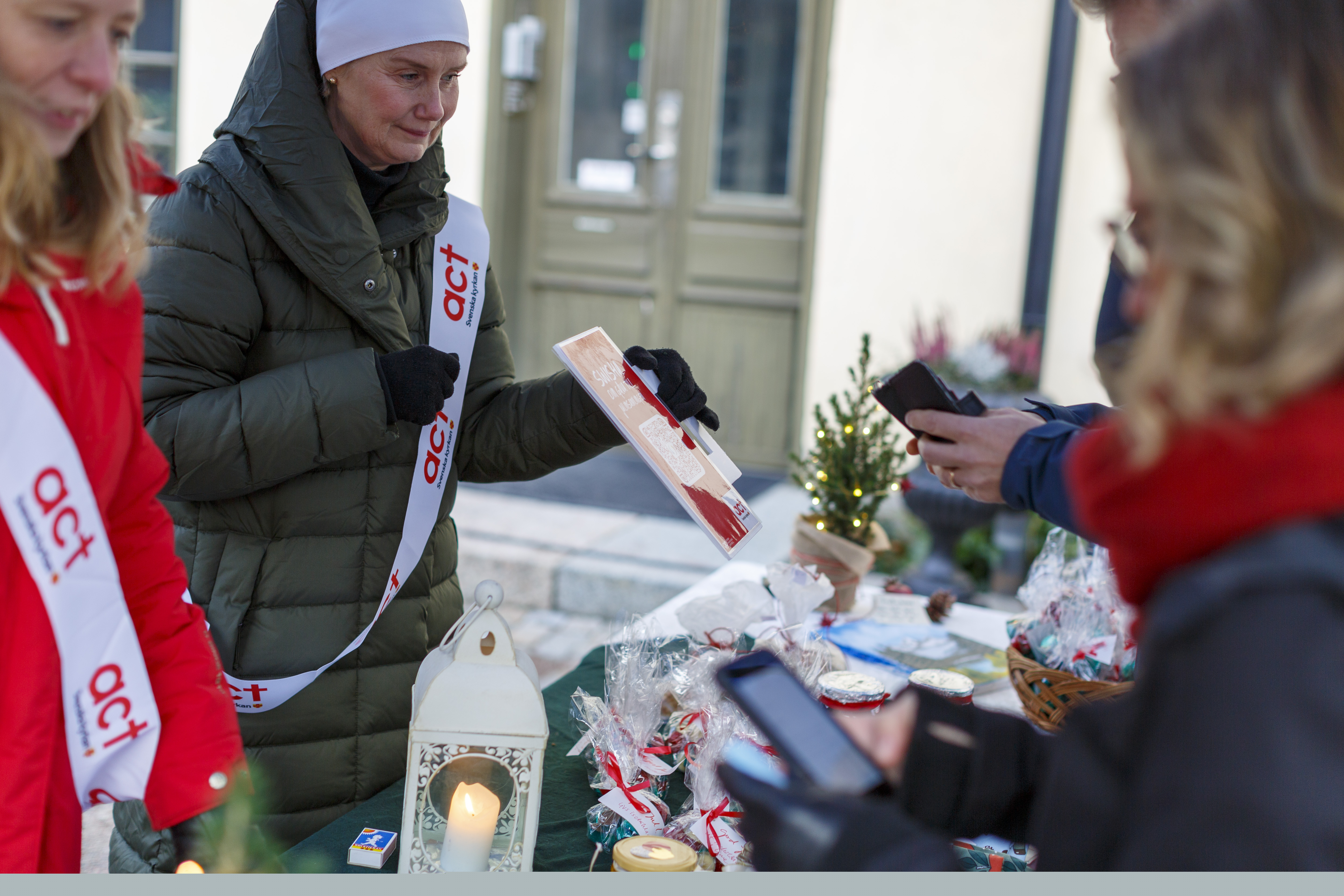 Två kvinnor med Act Svenska kyrkan banderoller över axeln säljer lotter och kakor på julmarknad.