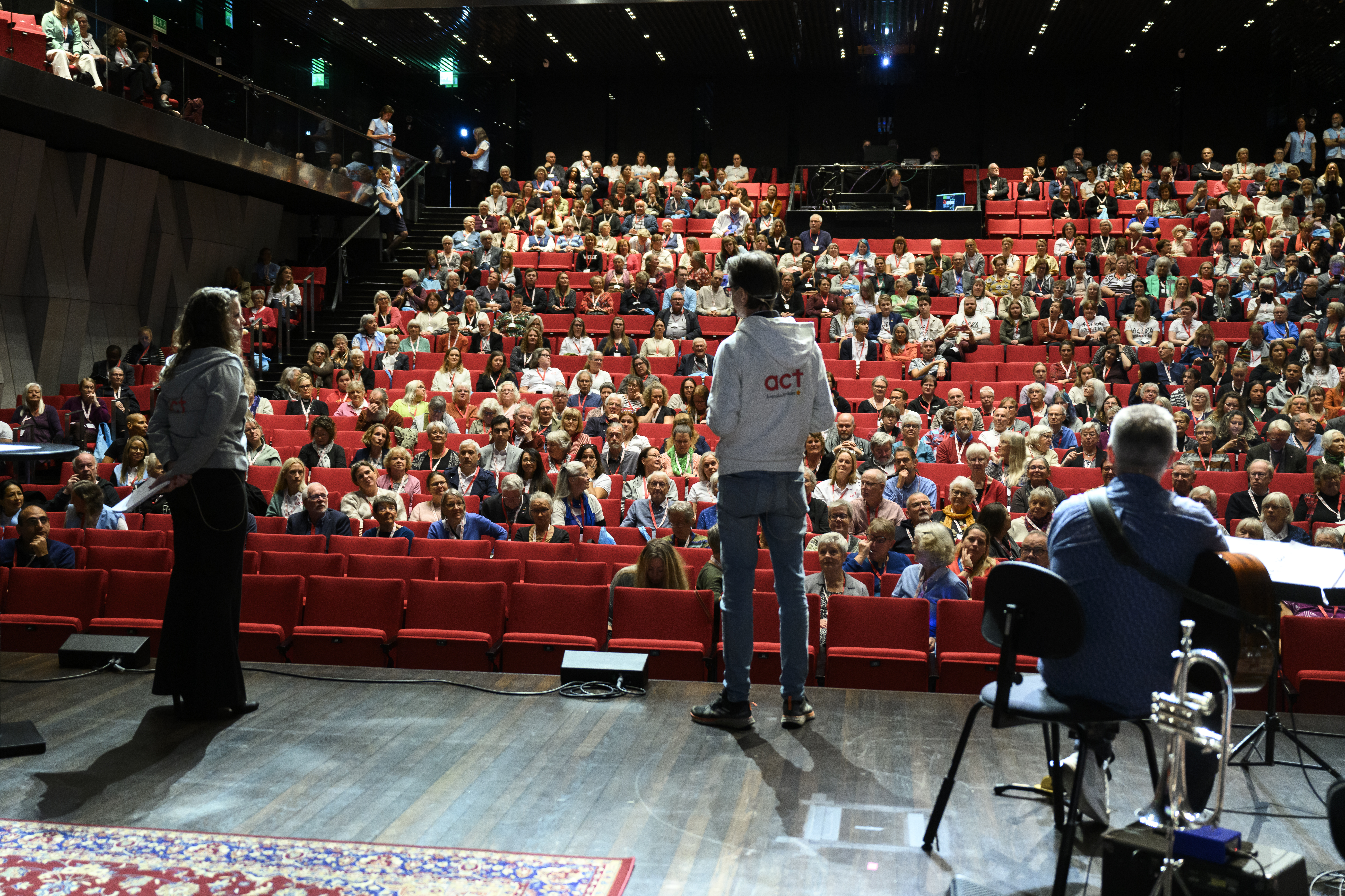 Ryggar på tre personer på en scen. I publiken ser vi flera hundra åskådare. 