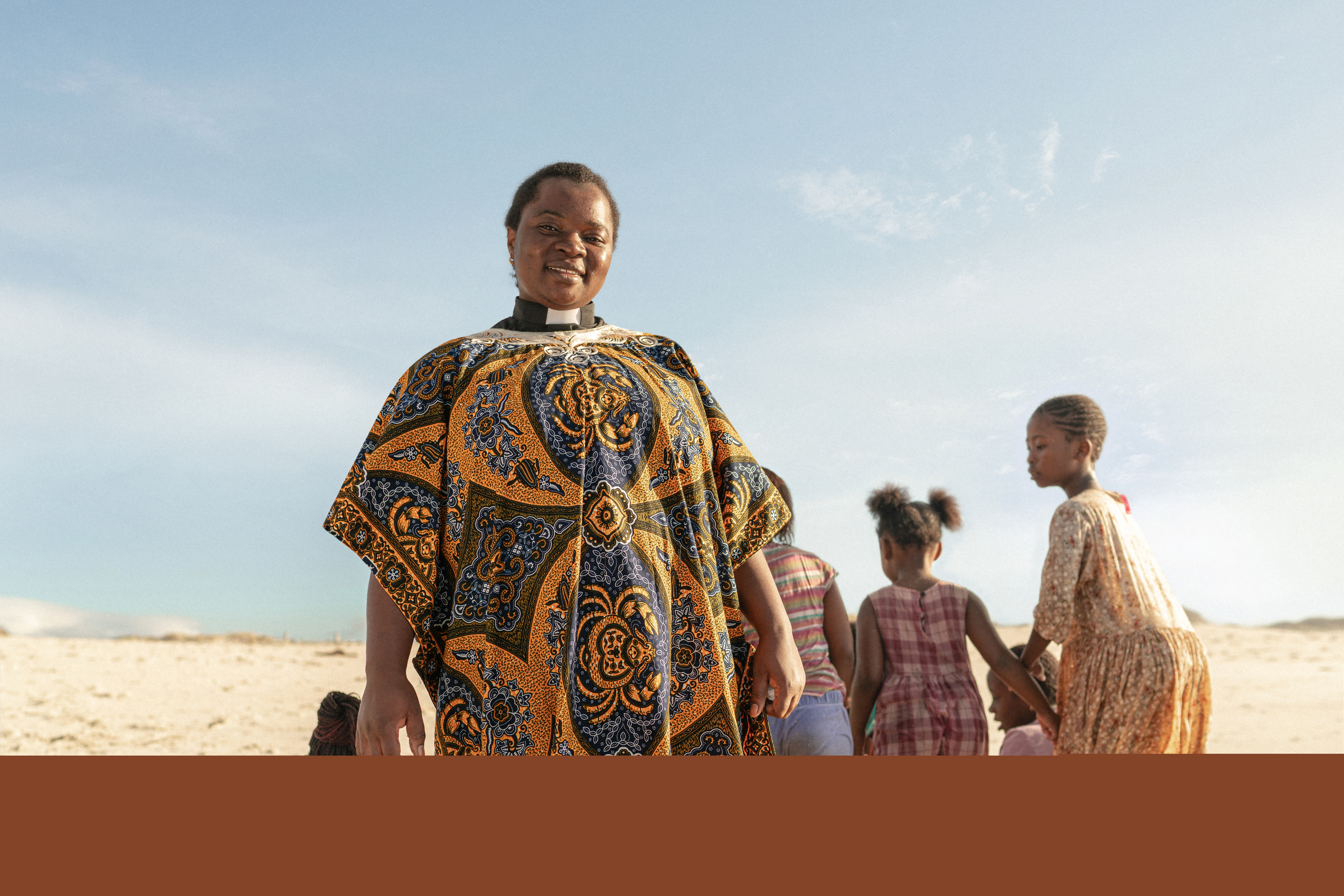 Kvinna i prästkläder på en strand. Bakom henne leker några flickor.