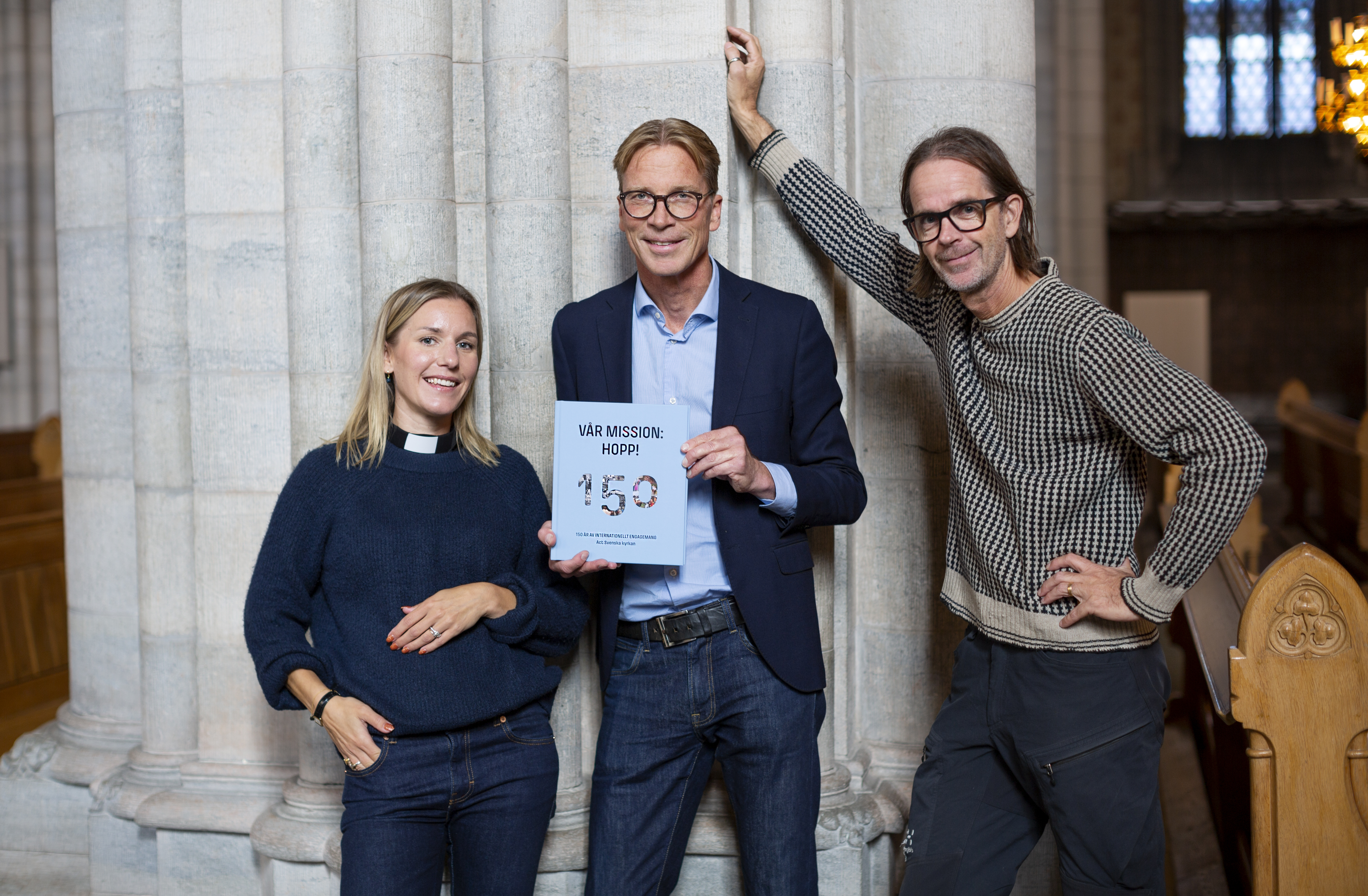 På bilden syns tre personer, personen i mitten håller i boken Vår mission: hopp!