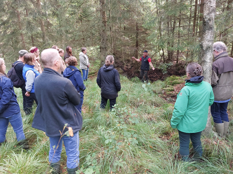 En grupp människor i stövlar och funktionsjackor är samlade i gräset invid ett dike, där en man står och berättar egnagerat.