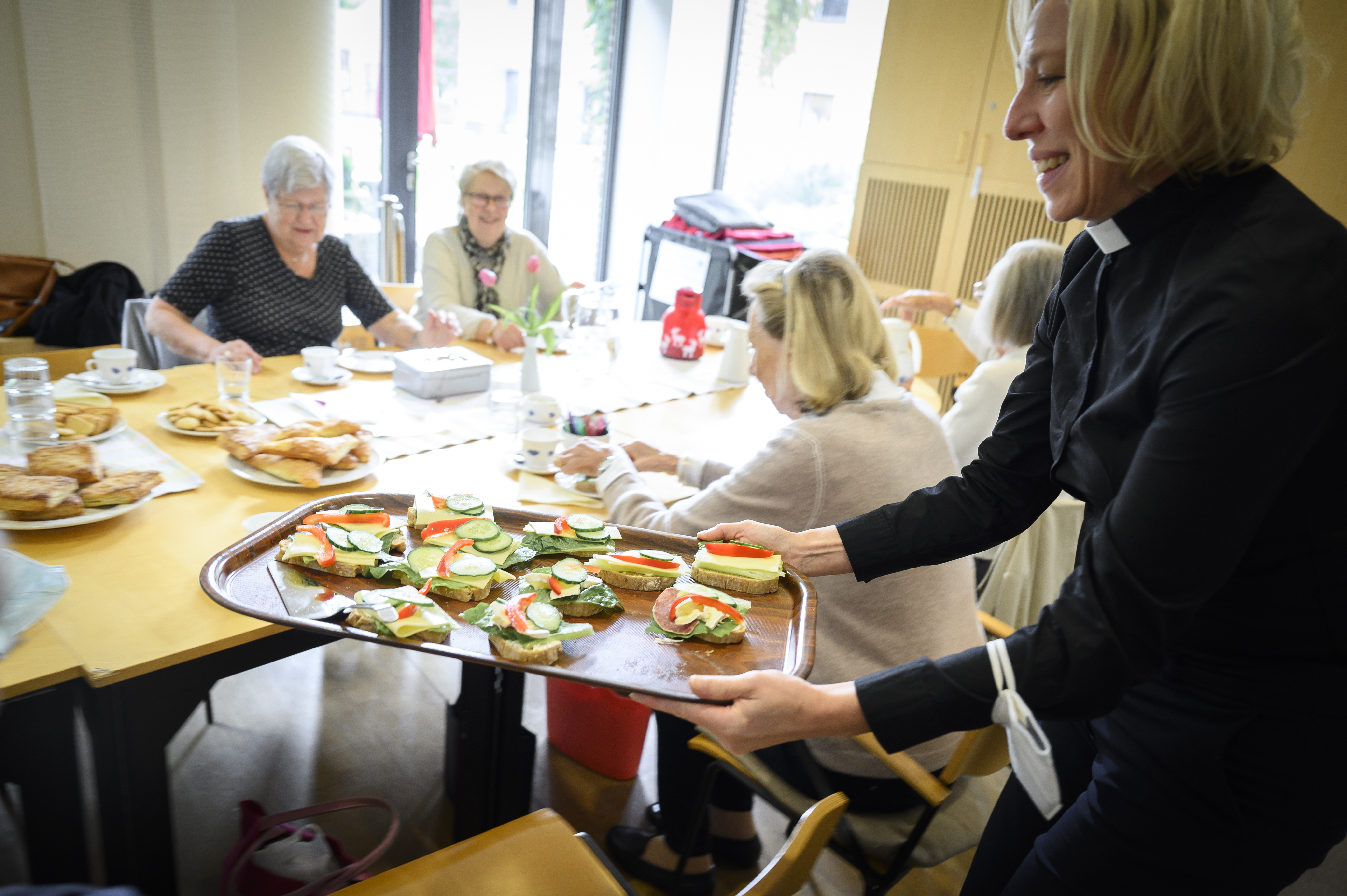 En kvinnlig präst delar ut smörgåsar till några damer som sitter runt ett bord.