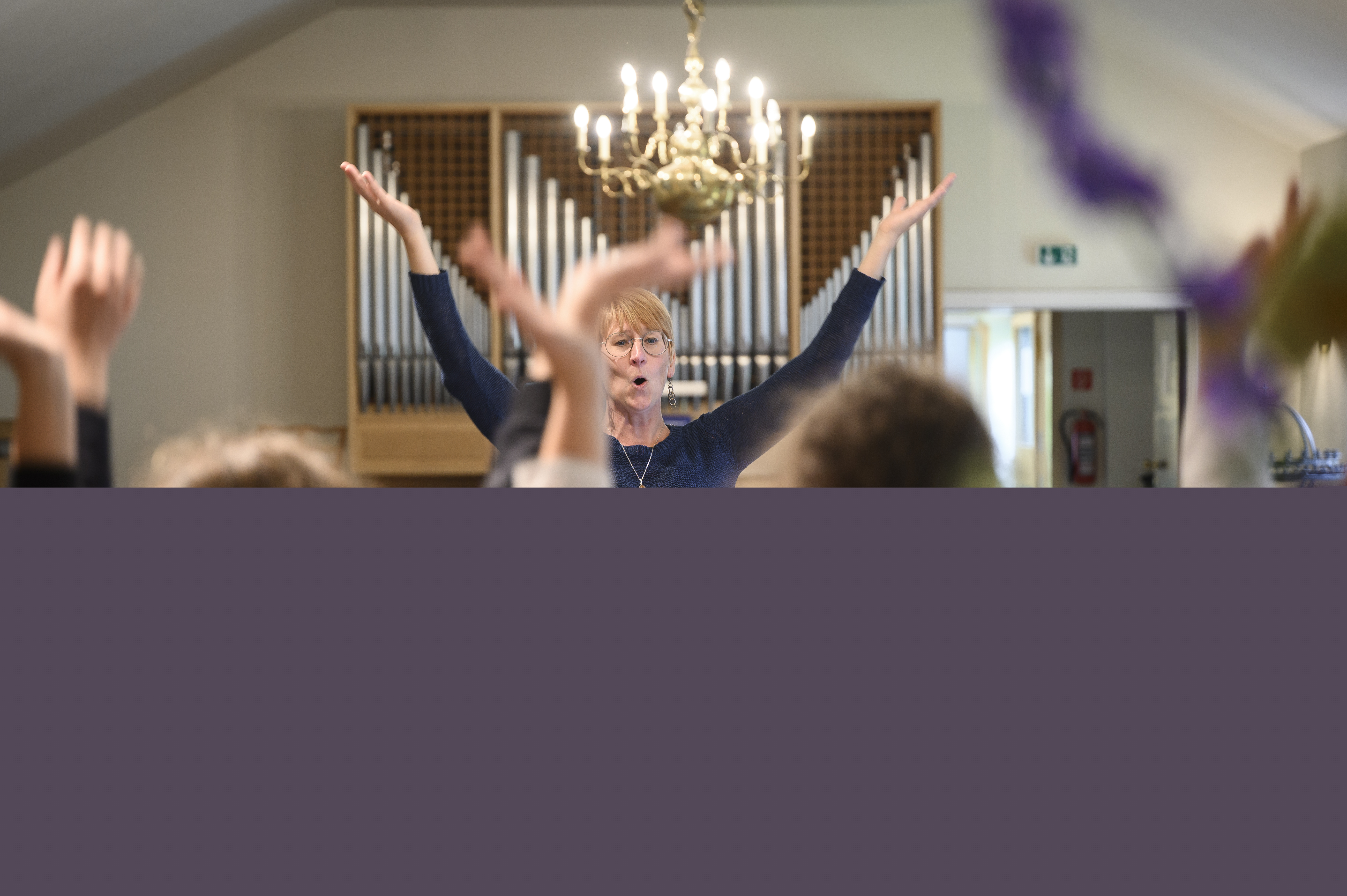 En kvinna står framför en orgel med armarna upp i luften. Några barn står i förgrunden och gör samma gest.